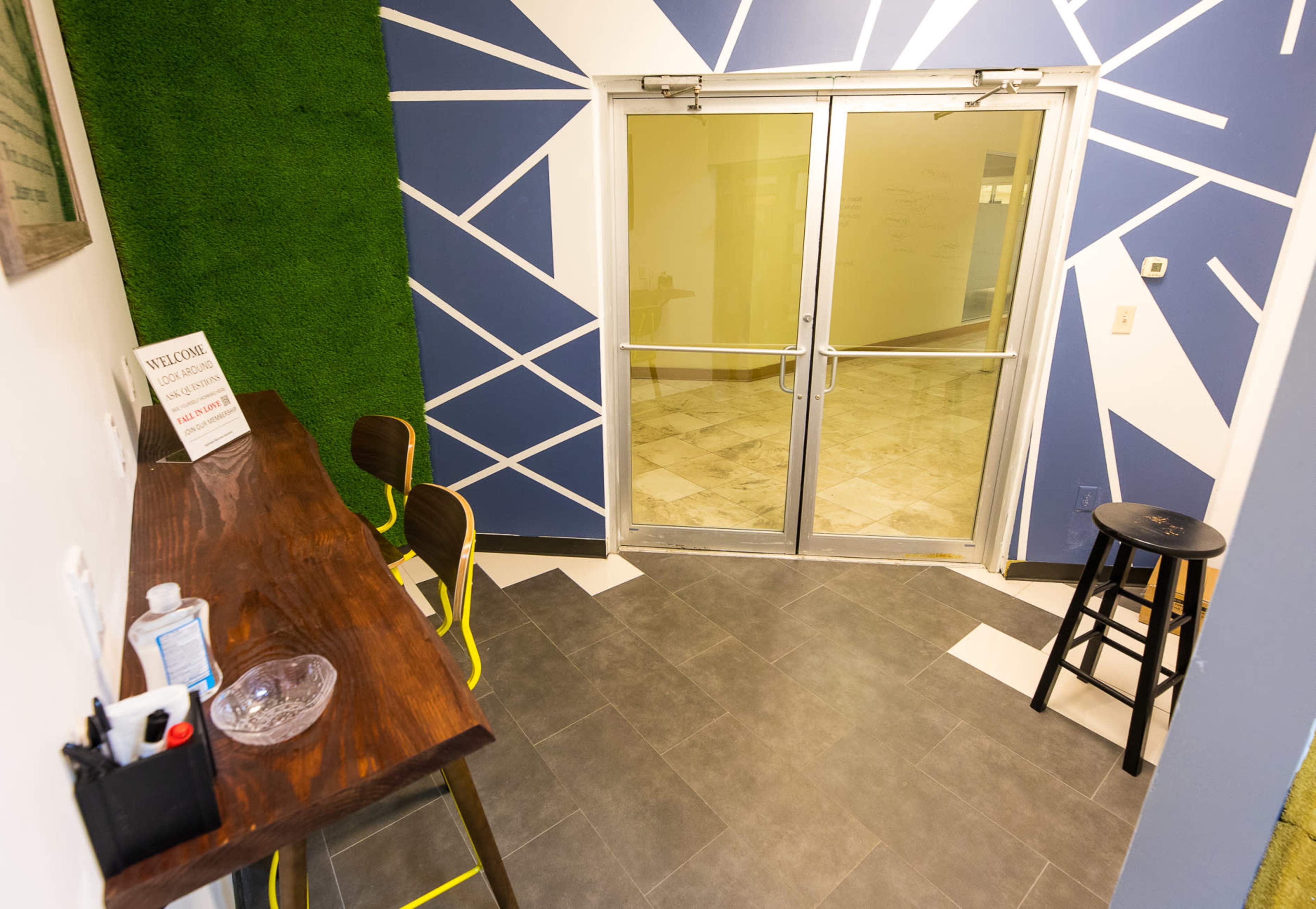 A modern entryway with a wooden counter, two chairs, and vibrant wall art, leading to glass double doors.