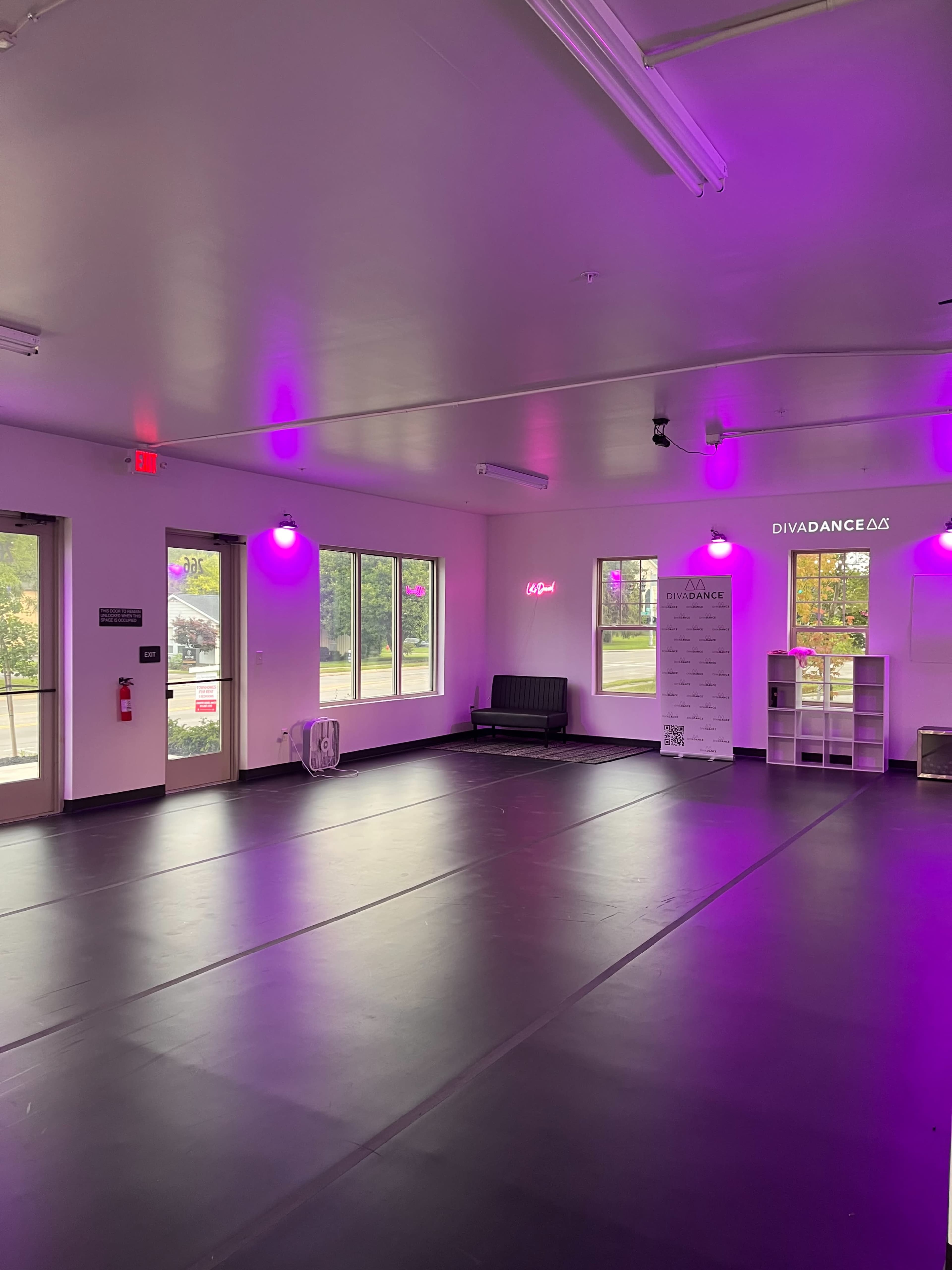 The interior of a dance studio features a black floor, pink lighting, and large windows with a view of the outdoors.