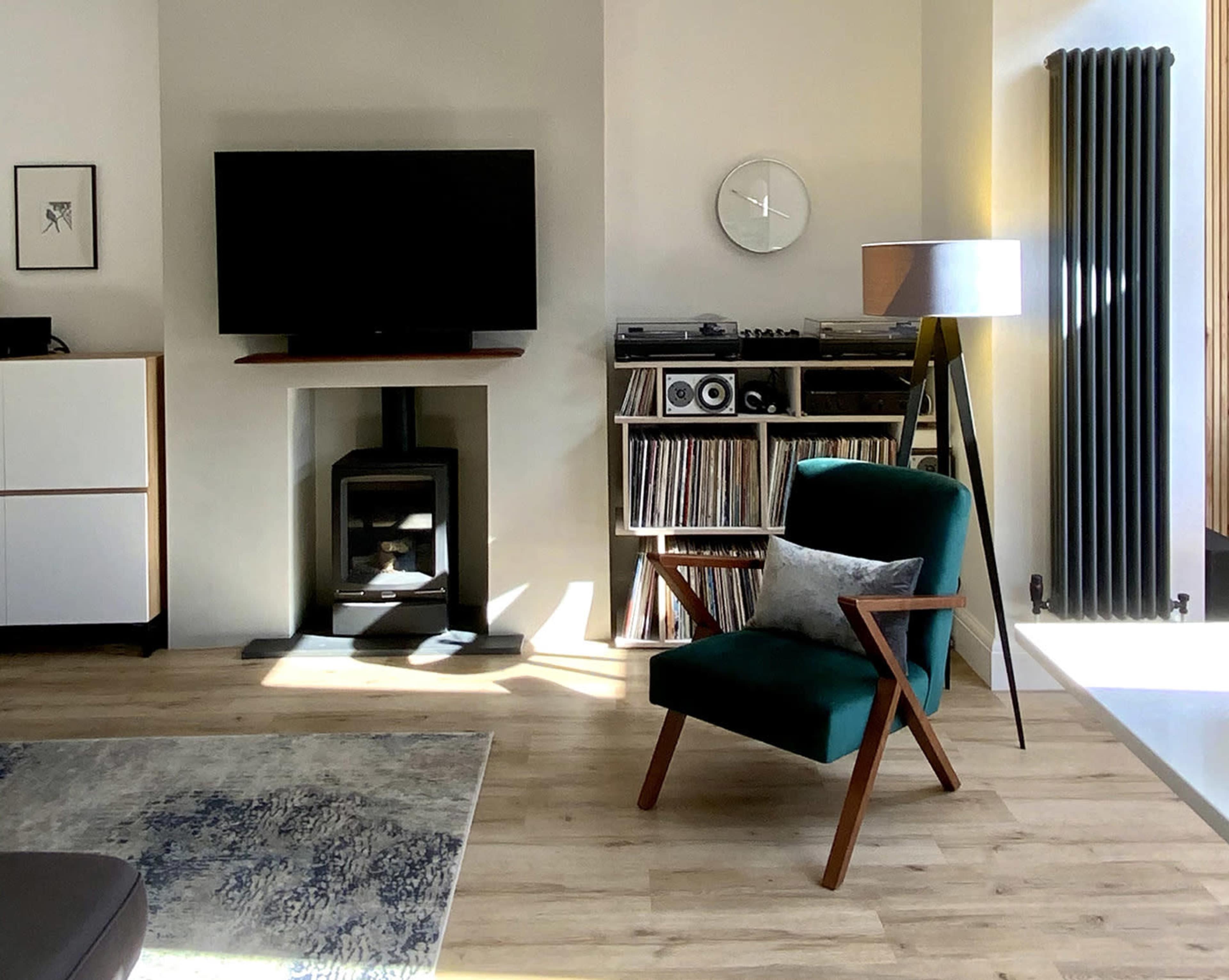 A modern living room with a green armchair, a television mounted on the wall, a wooden shelf displaying vinyl records, and a standing lamp beside a radiator.