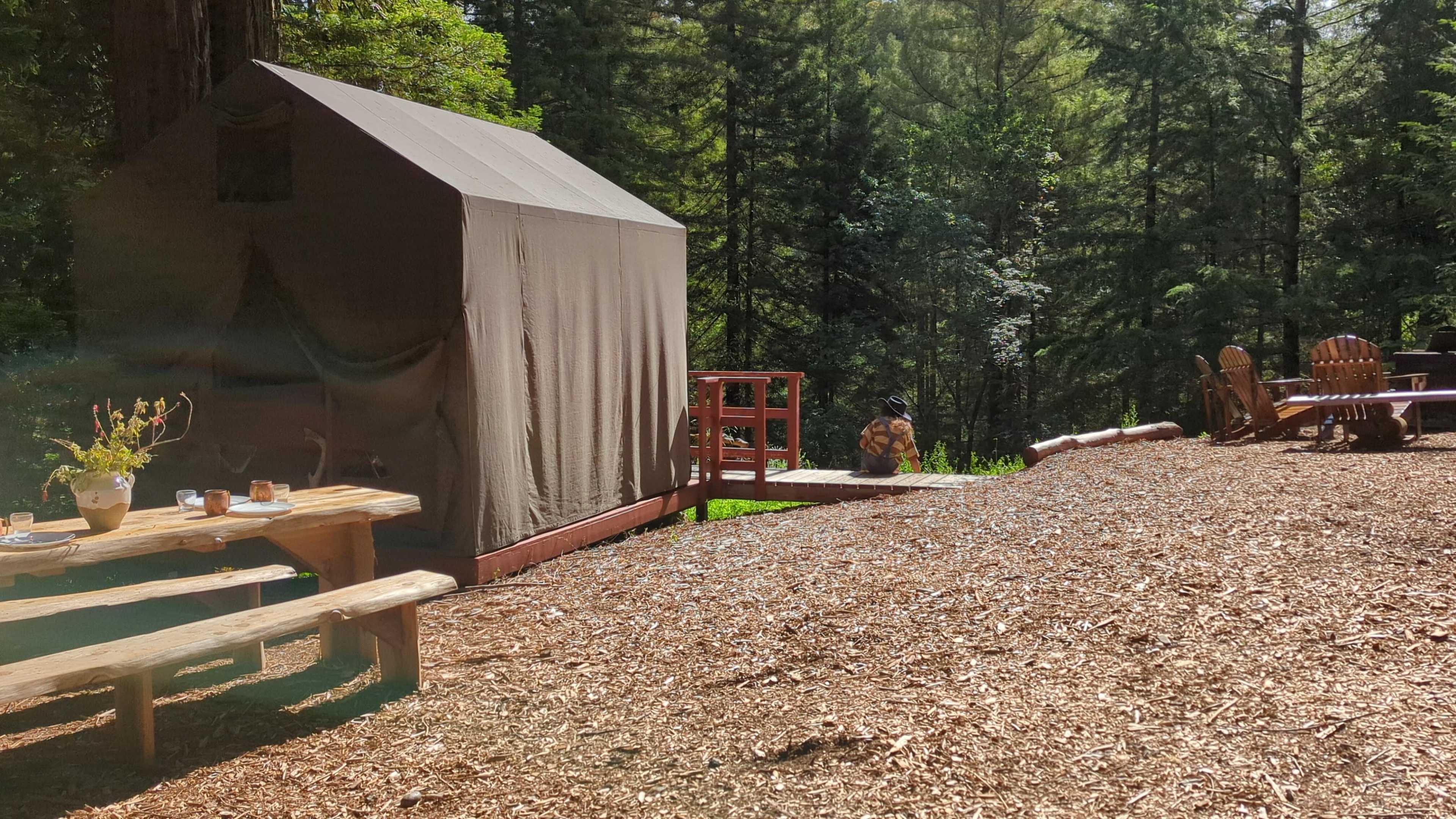 A tent sits on a wooden platform surrounded by trees, with a picnic table and several chairs nearby on a gravel surface.