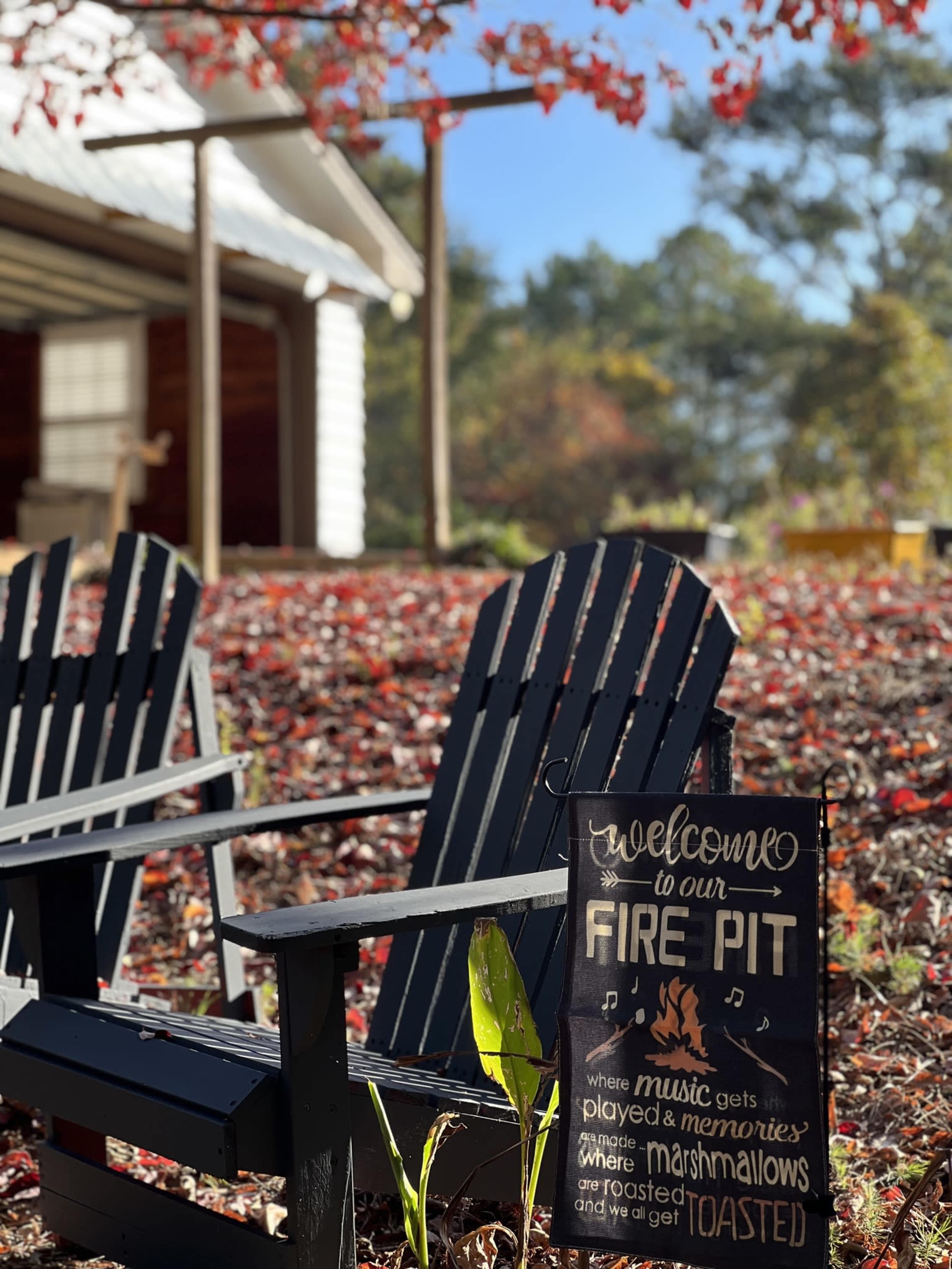 A sign welcoming guests to a fire pit area is positioned among fallen red leaves, with two black adirondack chairs visible nearby.