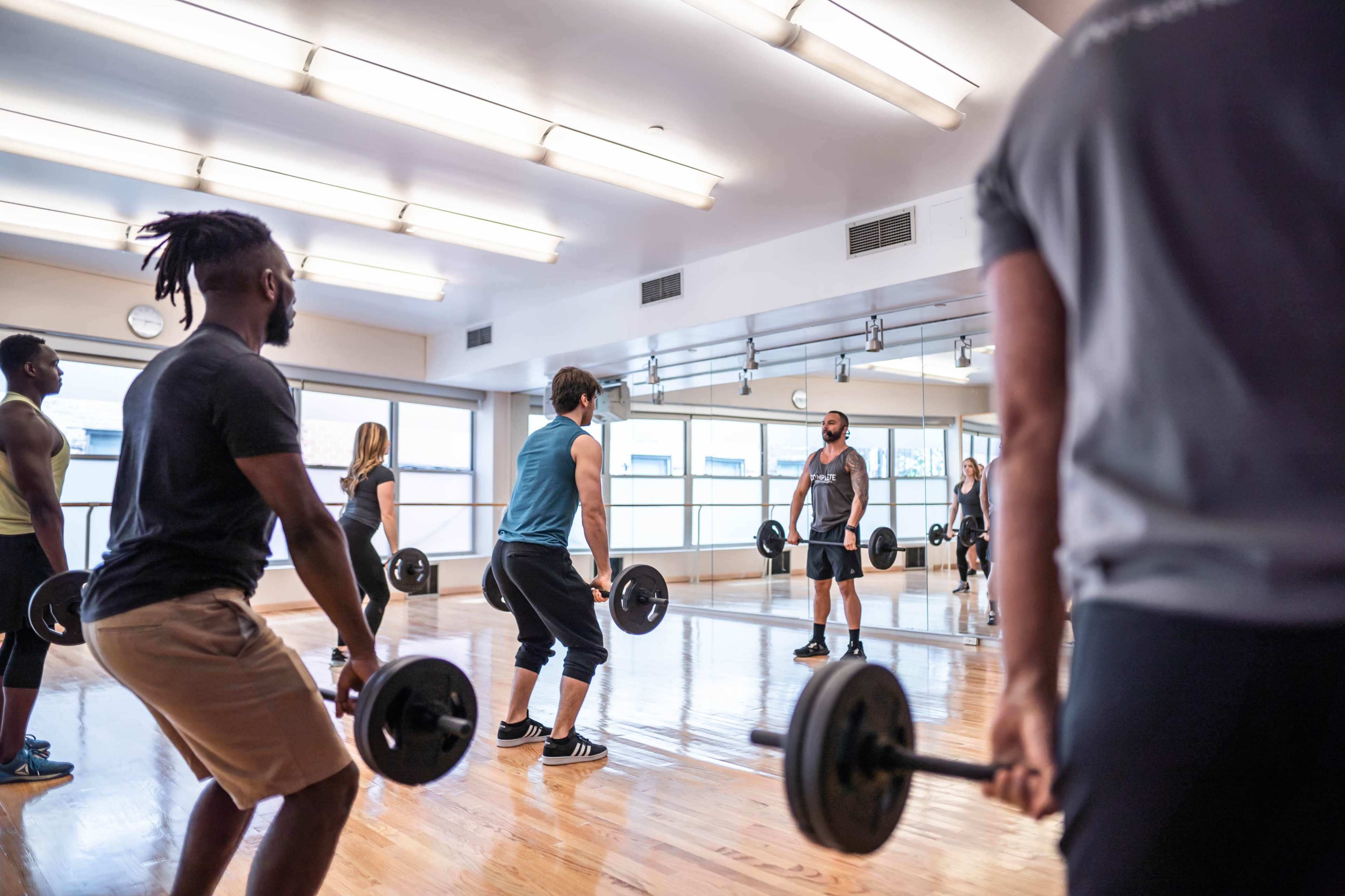 A group of people is lifting weights in a bright gym studio with mirrors and wooden floors.