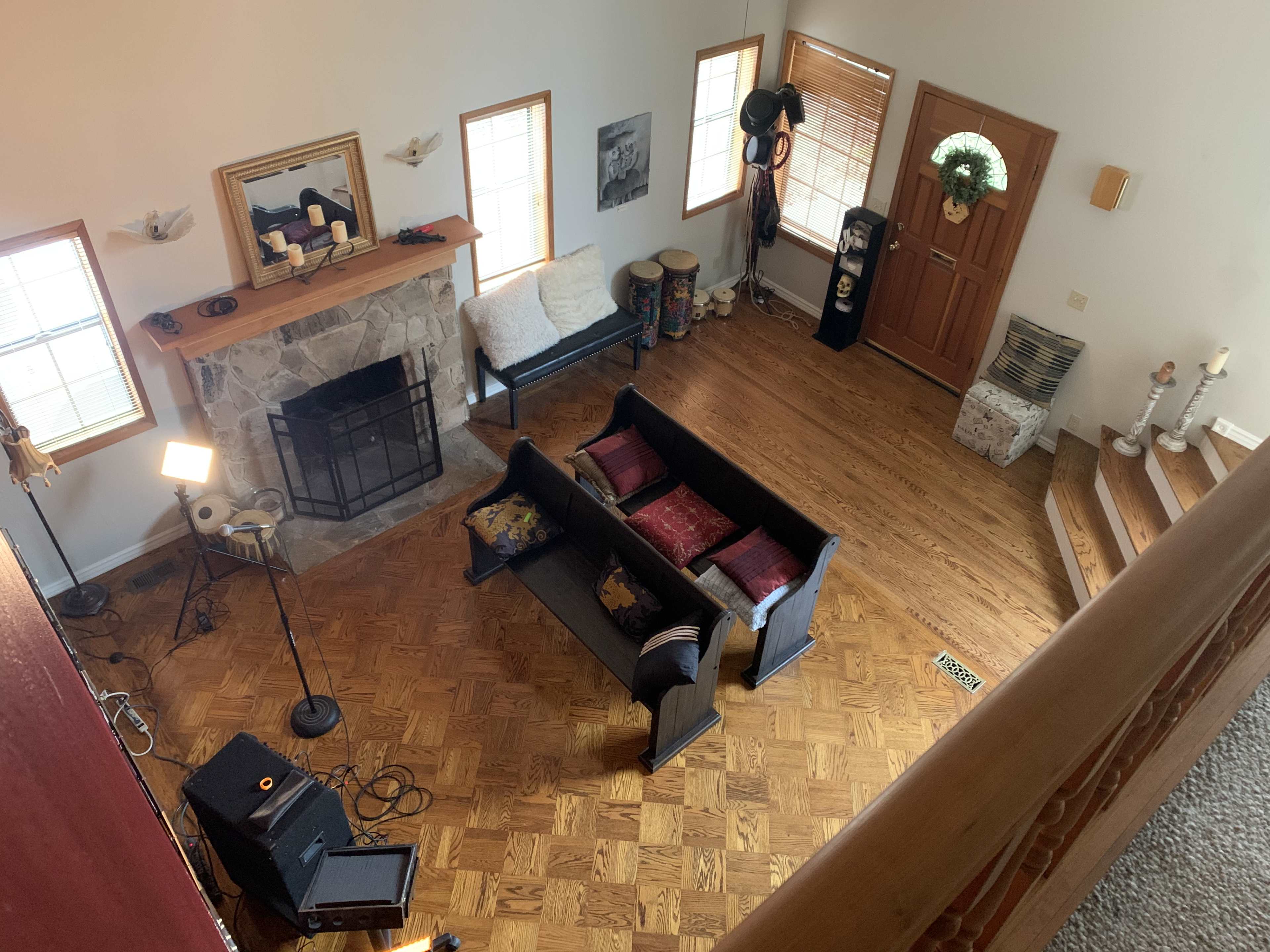 The image shows a two-story living room with a stone fireplace, a black couch facing a wooden door, and a staircase leading to an upper level.