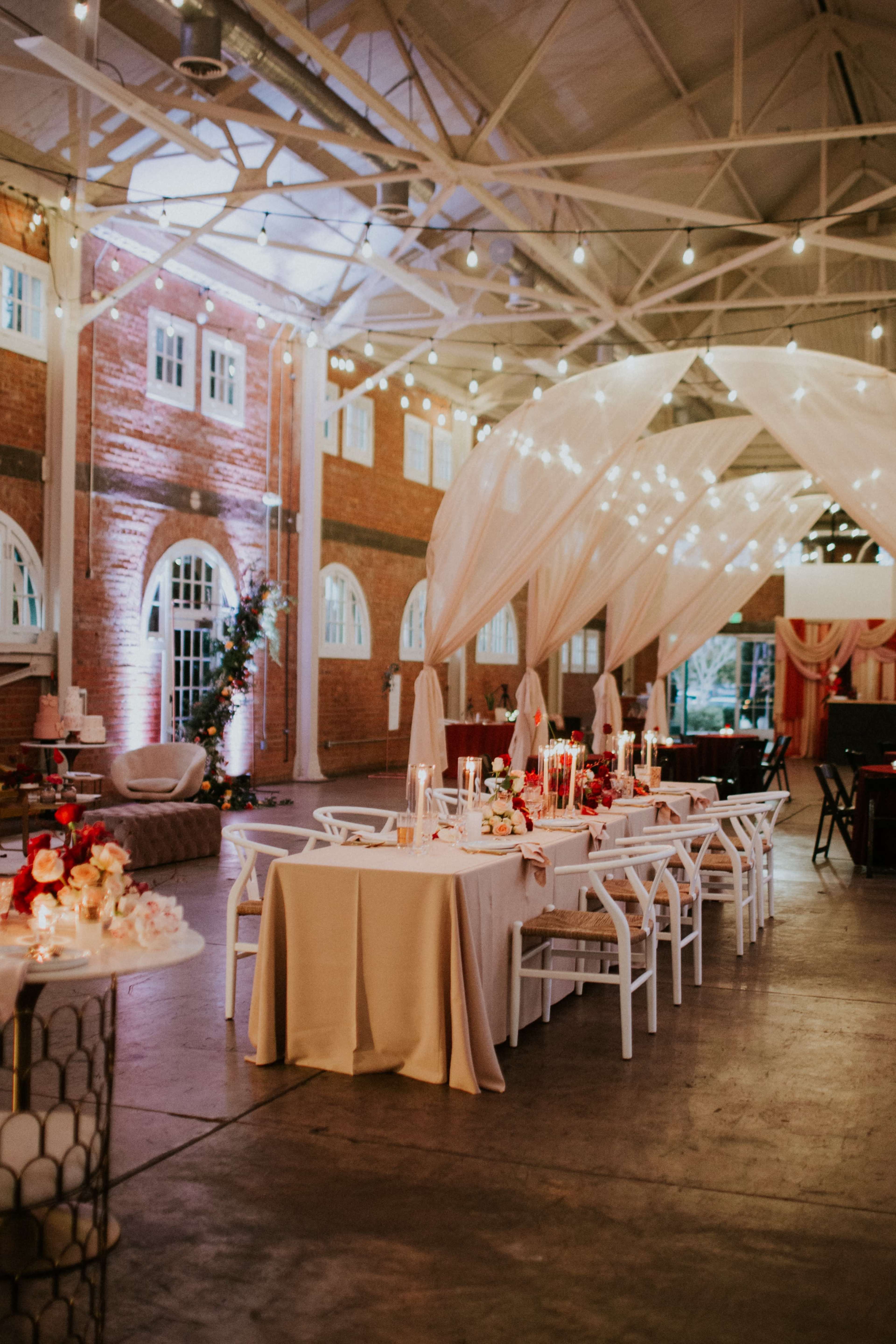 A spacious event hall features elegantly set tables with white chairs, draped fabric overhead, and string lights illuminating the area.
