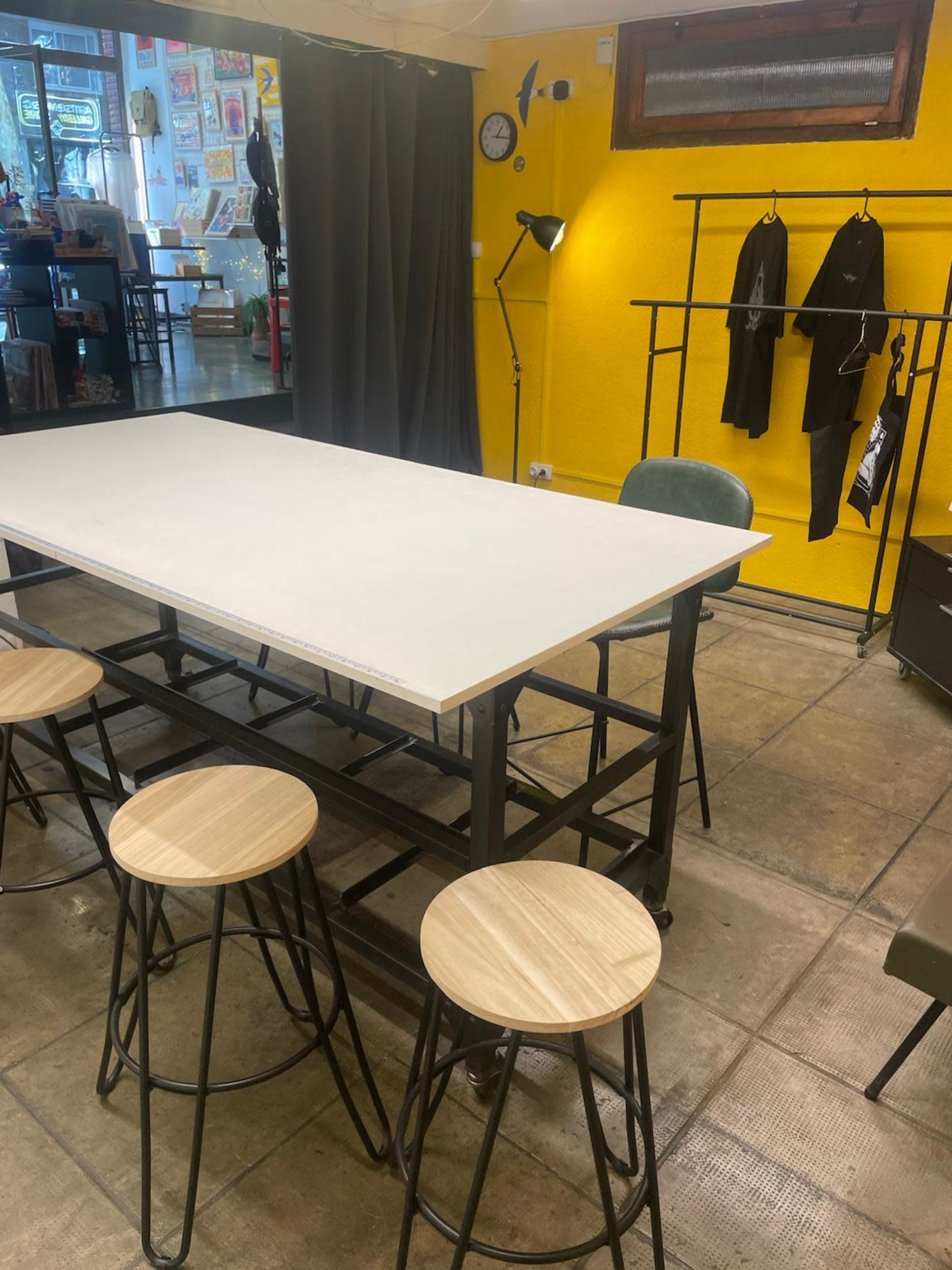 A large, rectangular table with wooden stools surrounds it in a brightly colored room featuring a yellow wall, shelves, and clothing racks.