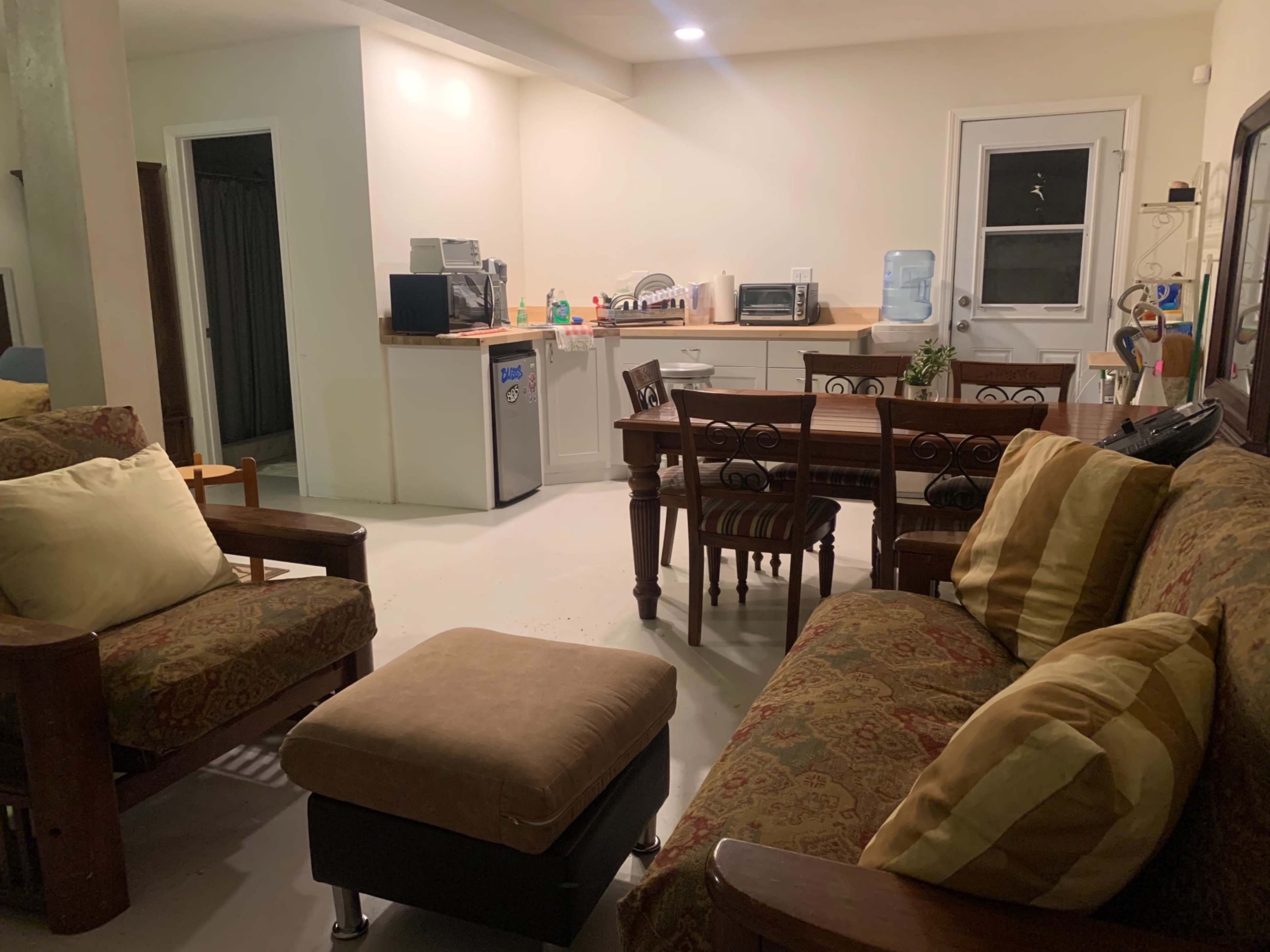 A living area with two couches, an ottoman, and a dining table near a kitchenette with a refrigerator and various appliances.