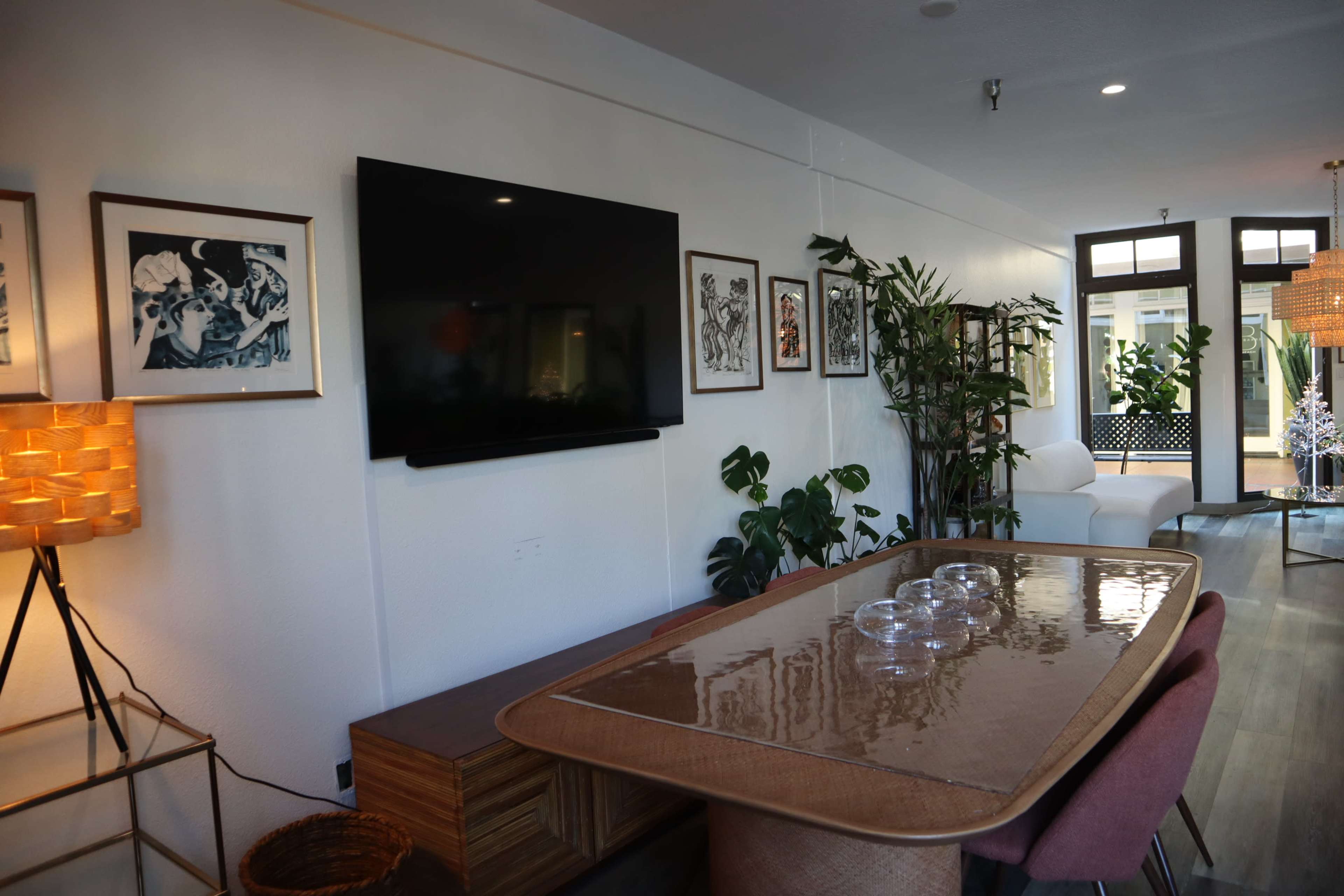 A modern living space featuring a dining table, a flat-screen TV on the wall, and an arrangement of plants and artwork.