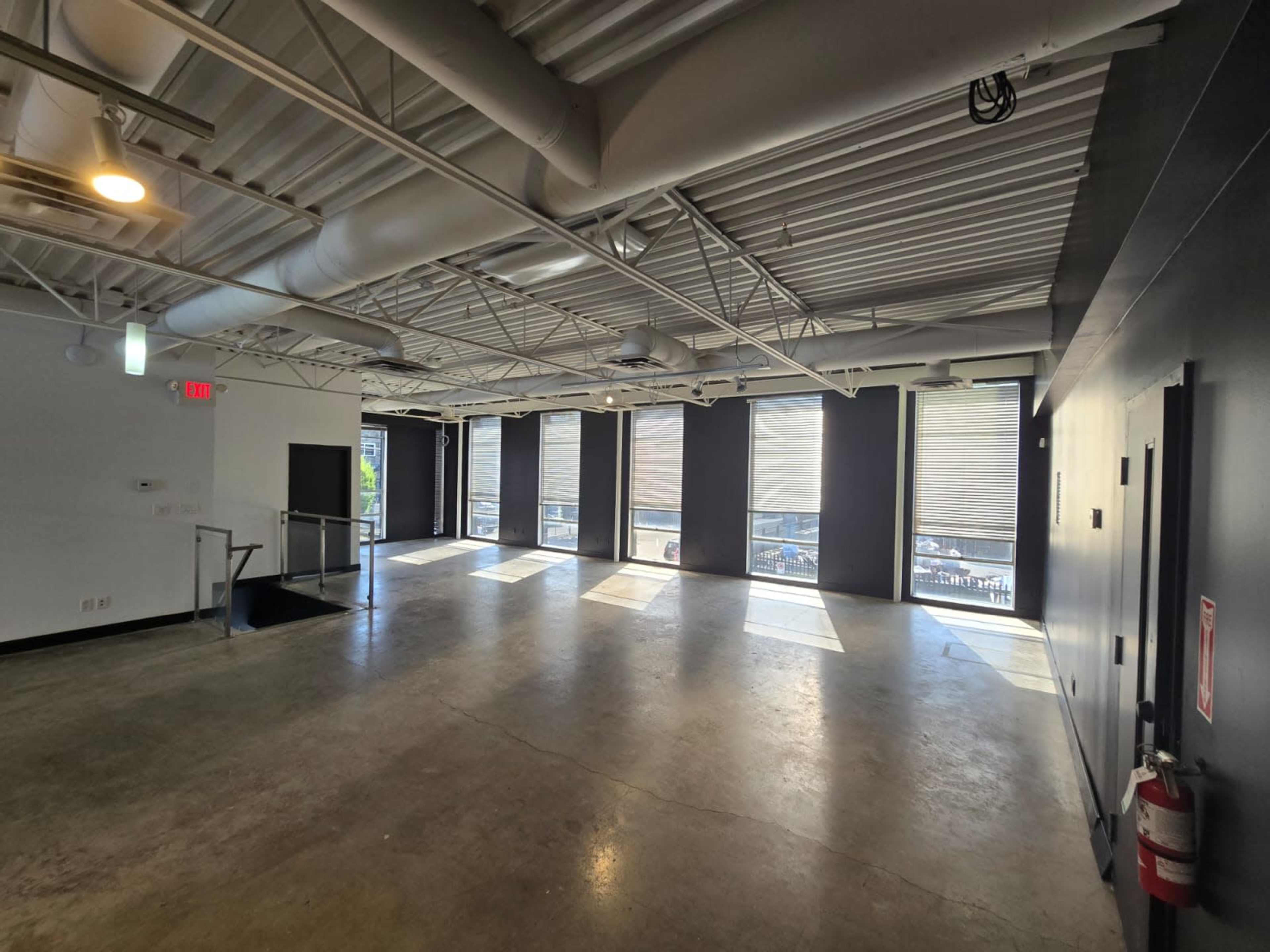 The image shows a spacious, empty room with large windows allowing natural light, and concrete flooring.