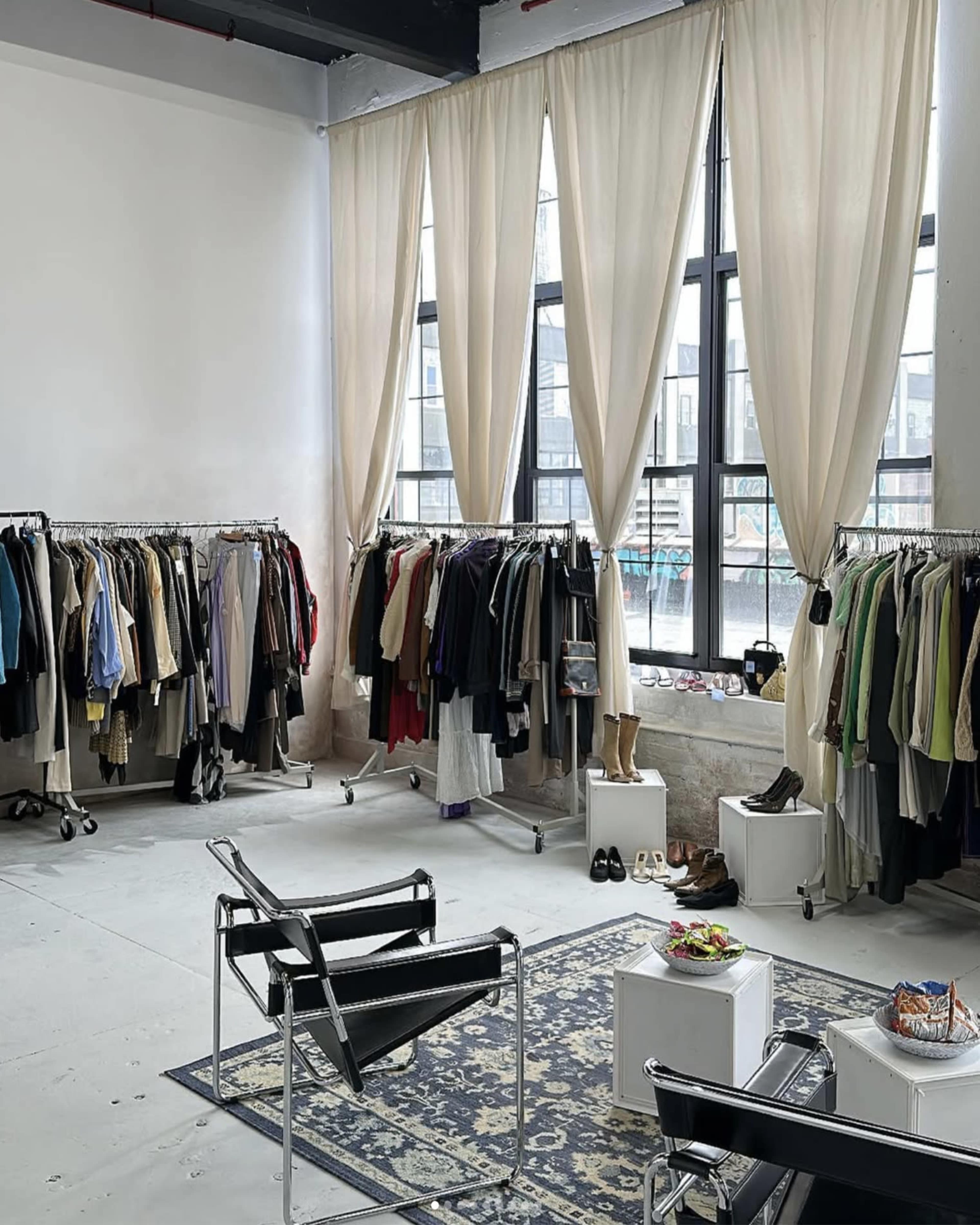 The image shows a spacious room with large windows, featuring clothing racks filled with various garments, a couple of chairs, and a small table with snacks.