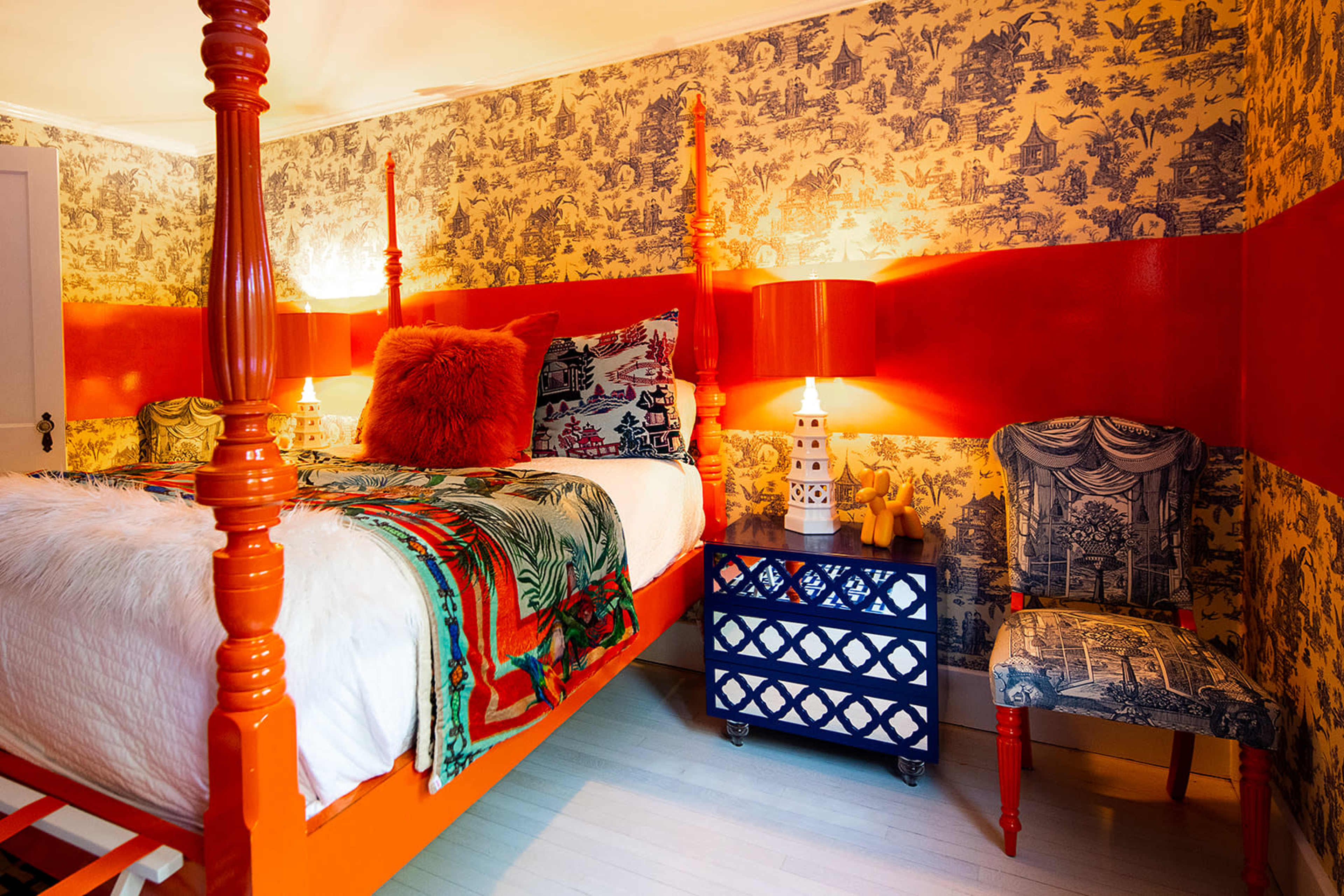 A brightly colored bedroom features a four-poster bed with an orange frame, decorative pillows, and a patterned blue chair beside a small blue nightstand.