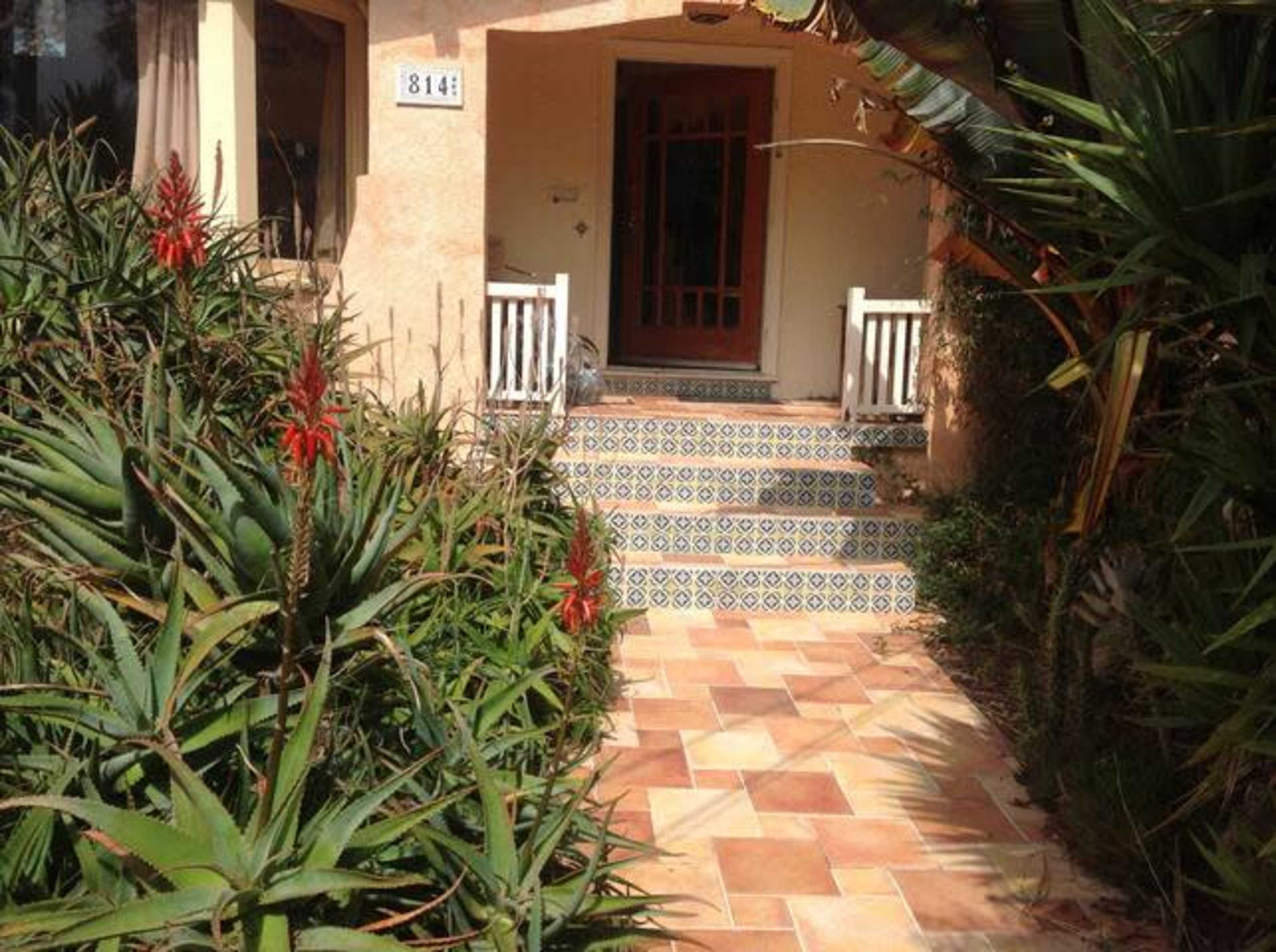 A front entrance of a house with a tiled pathway and surrounding greenery featuring red flowers.