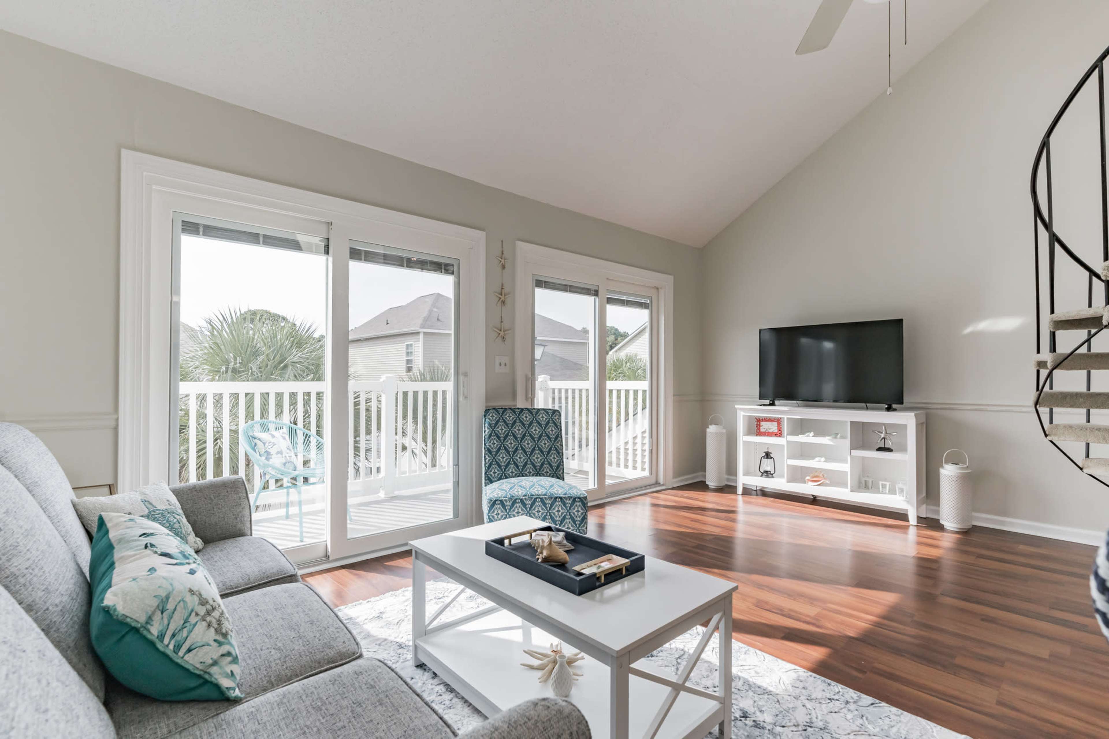 A bright living room features a gray sofa, a white coffee table, and a TV stand, with large windows opening to a balcony.