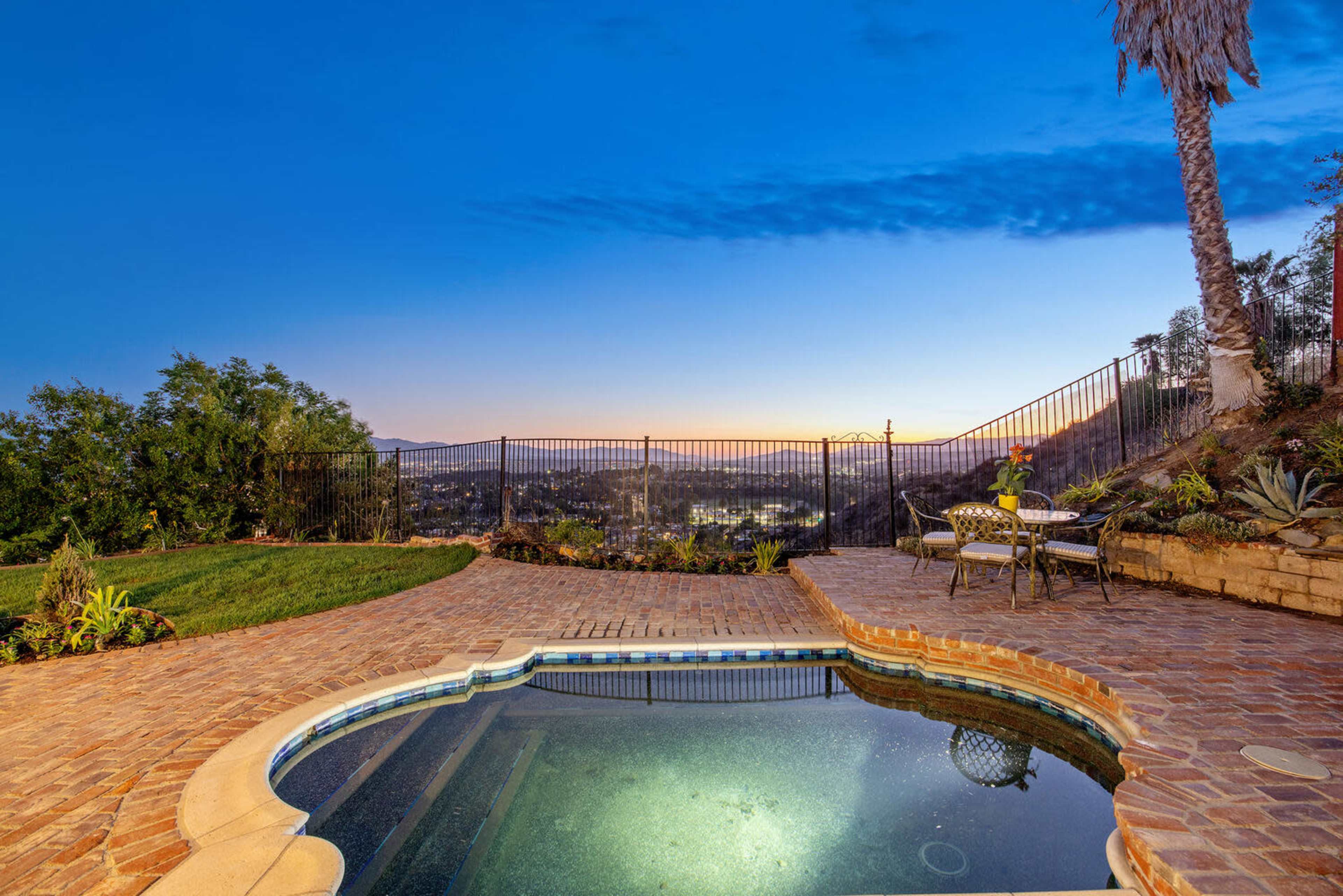 The image shows a backyard with a swimming pool, patio area, and a view of a valley at dusk, surrounded by trees and a fence.