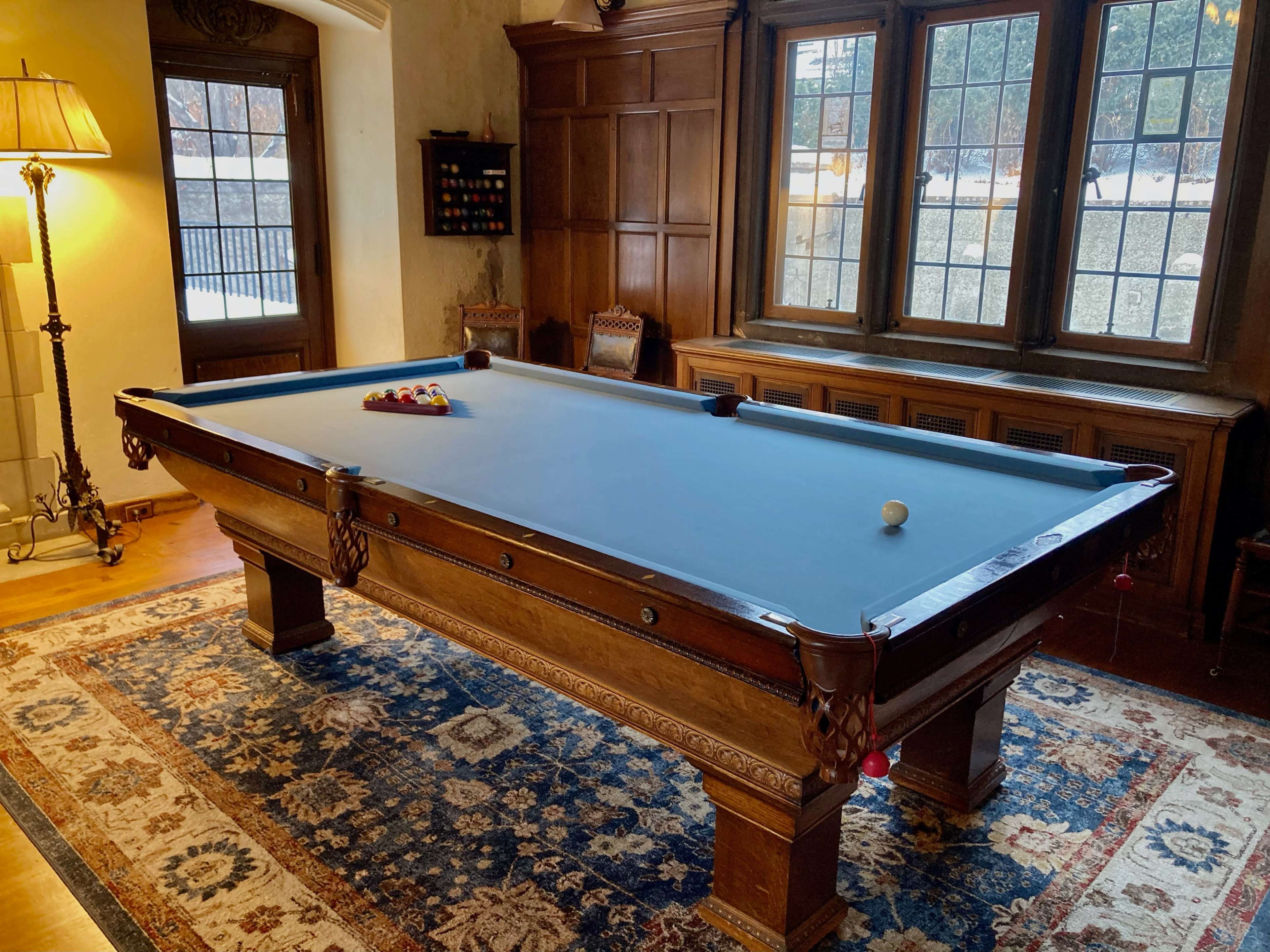 A wooden billiard table with a blue felt surface is positioned in a room featuring large windows and warm wood paneling.