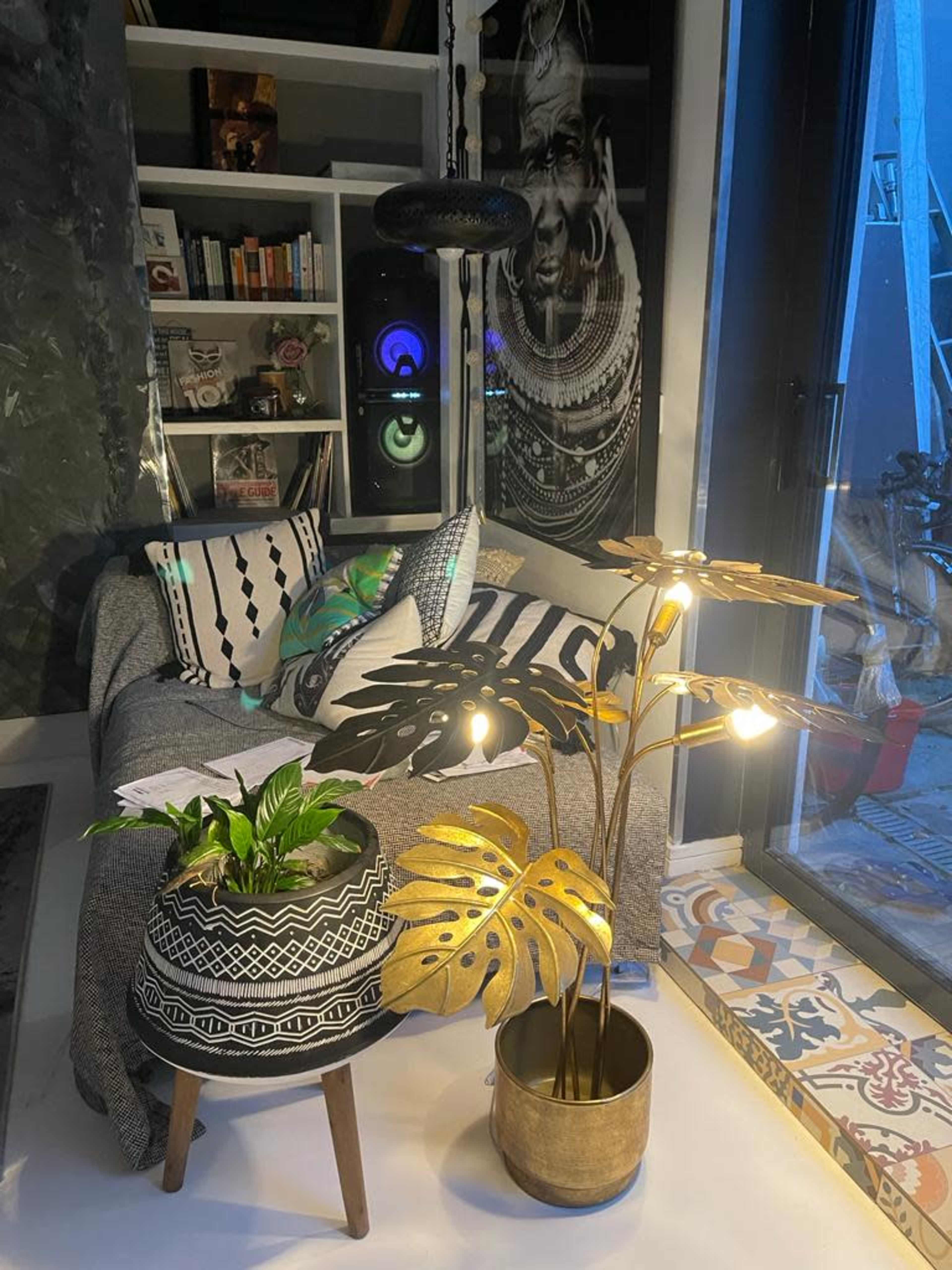 A modern living room features a grey couch adorned with patterned pillows, a plant in a decorative pot, and a lamp with leaf-shaped fixtures, all set against a backdrop of patterned tiles and shelves.