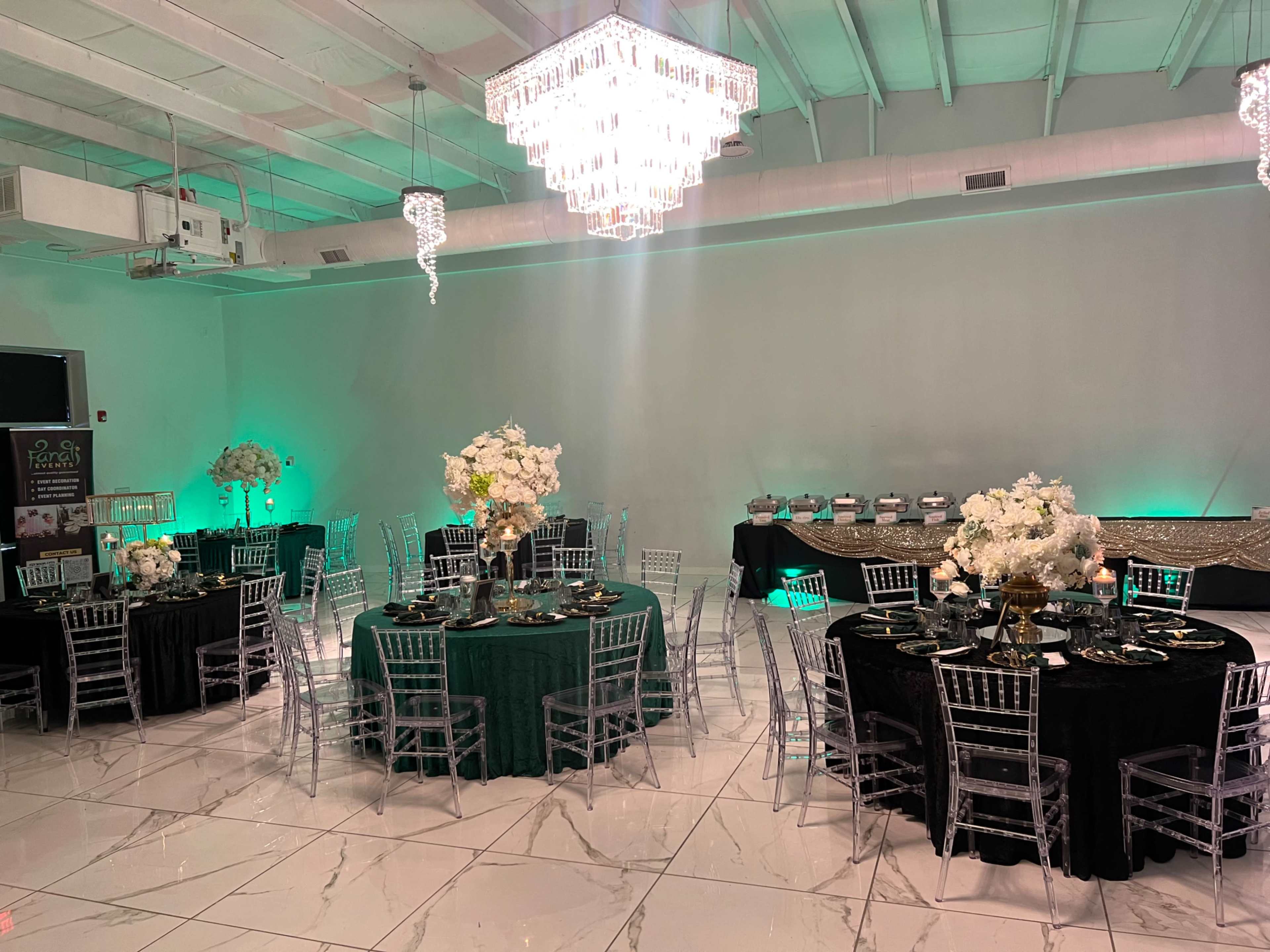 The image shows a banquet hall set up with round tables covered in black and green linens, adorned with floral centerpieces, and illuminated by a large chandelier and green lighting.