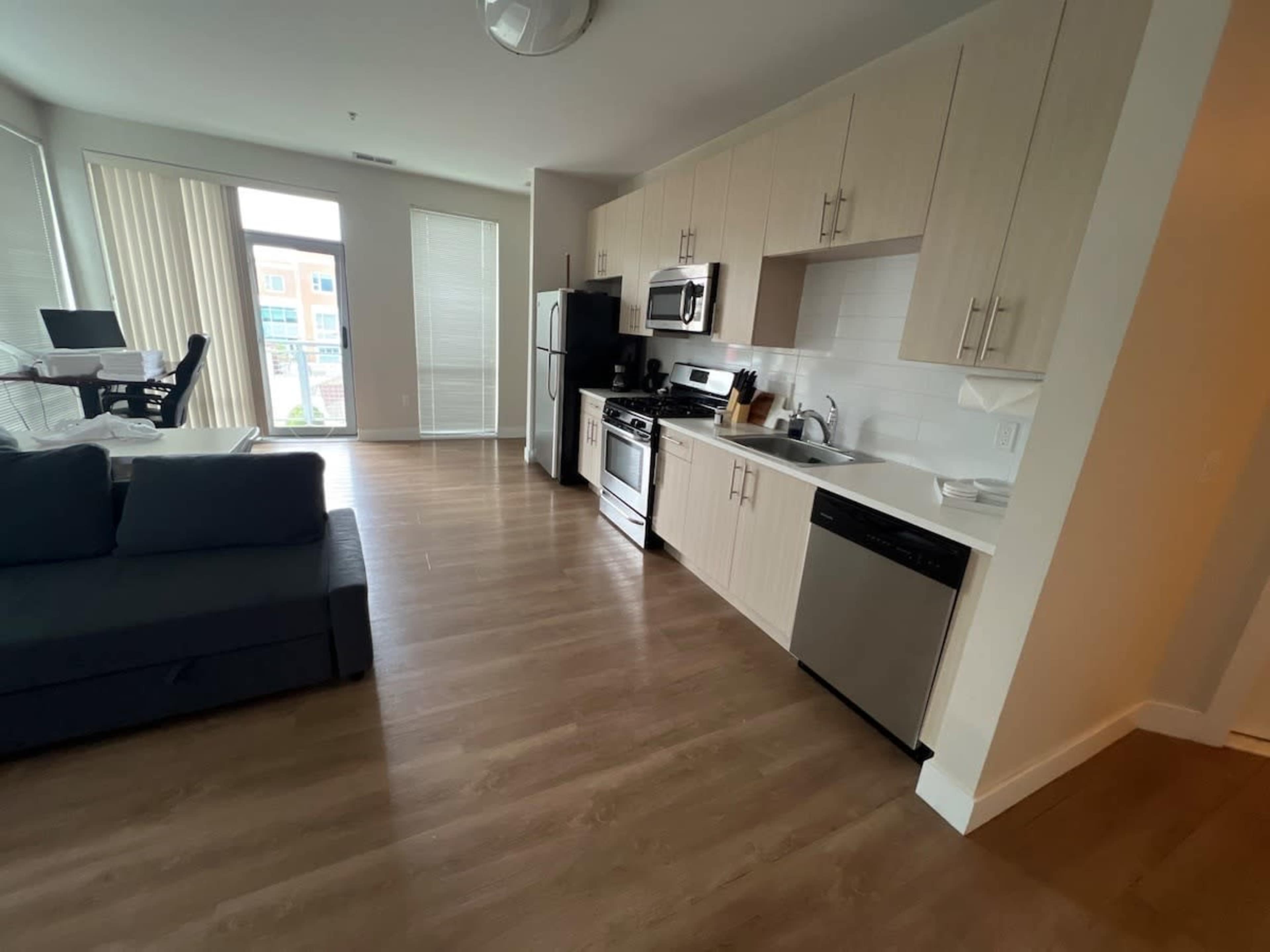 The image shows a modern kitchen and living area in an apartment, featuring light-colored cabinets, stainless steel appliances, and a sectional sofa.