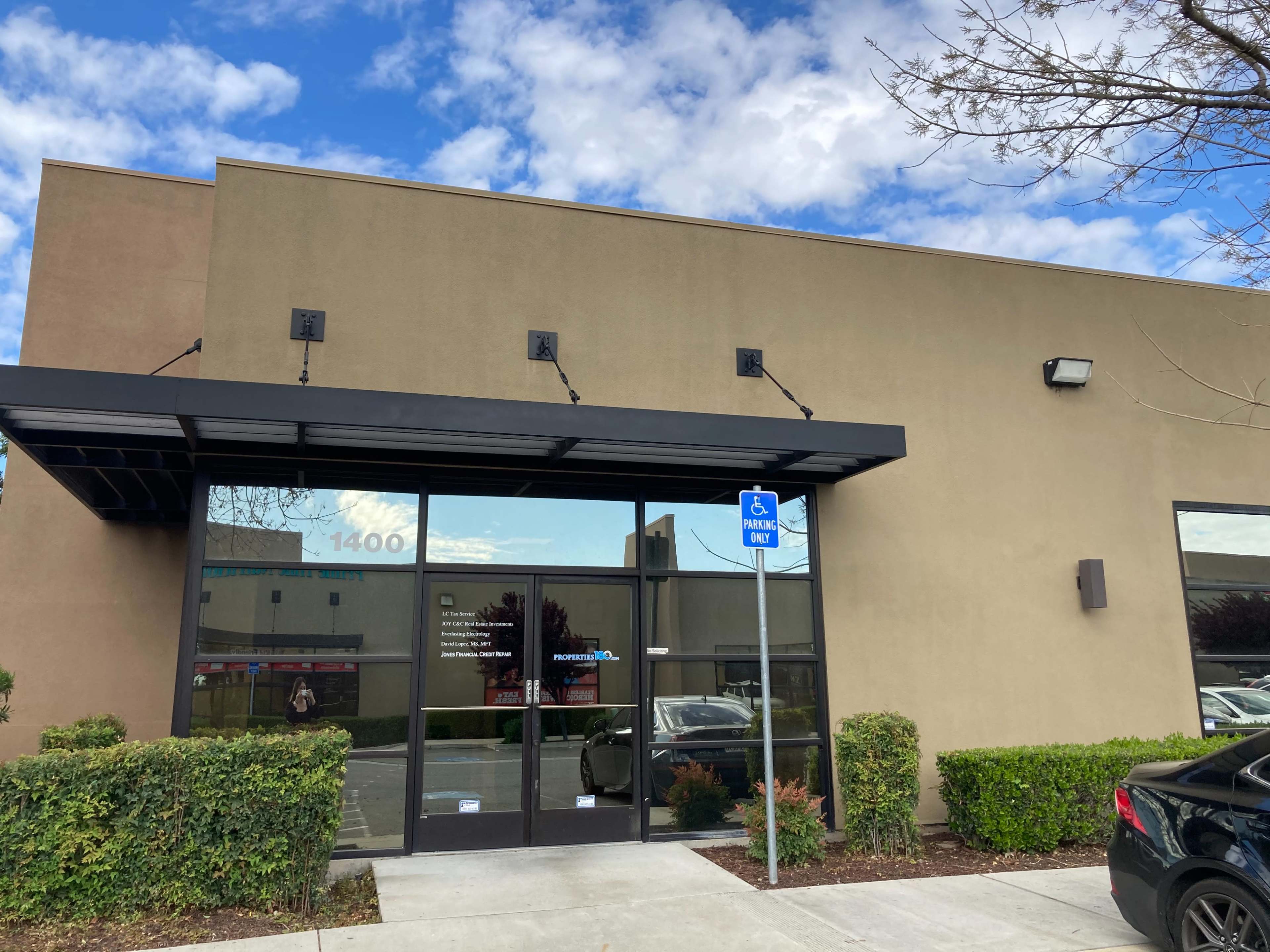 A single-story building features a glass entrance with a black overhang and a concrete sidewalk alongside a visible parking sign.