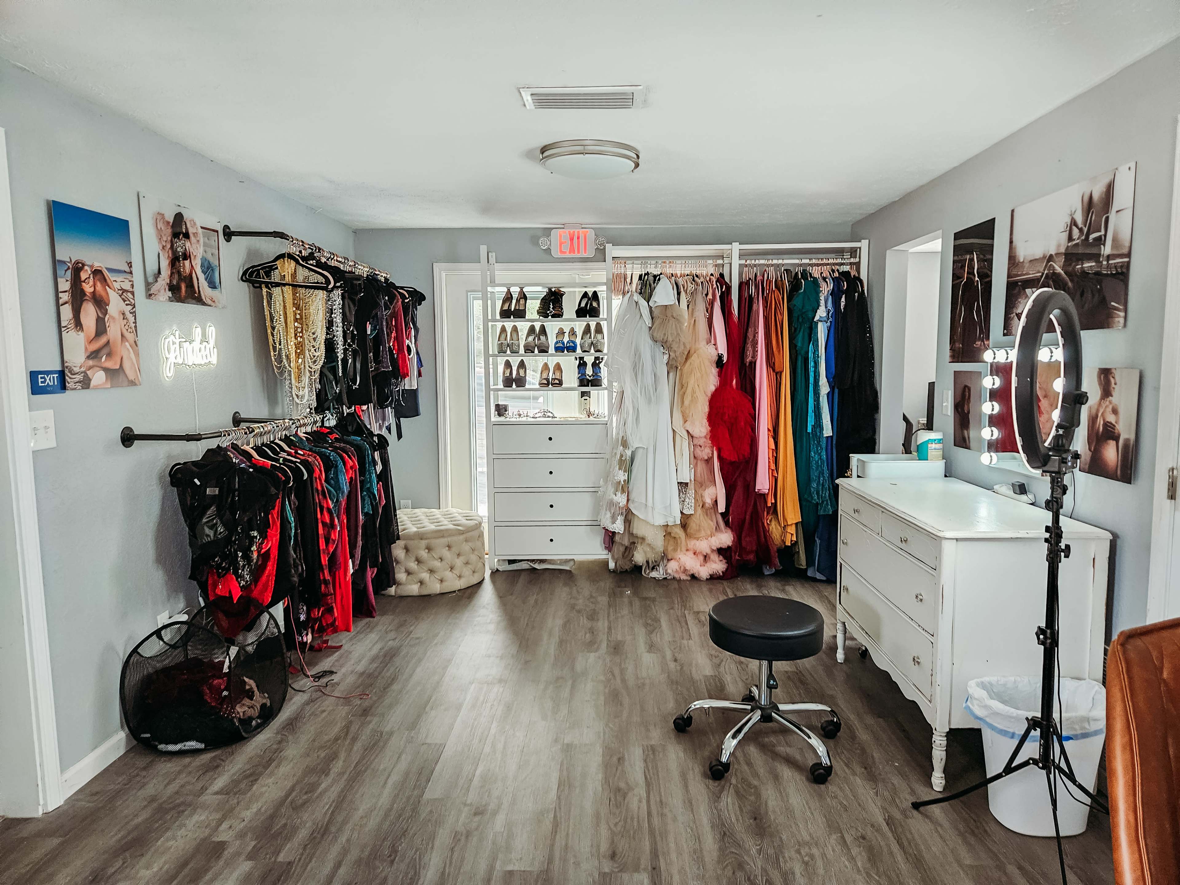 A well-organized dressing room with racks of colorful clothing, a white dresser, a vanity with a ring light, and several pairs of shoes displayed on shelves.