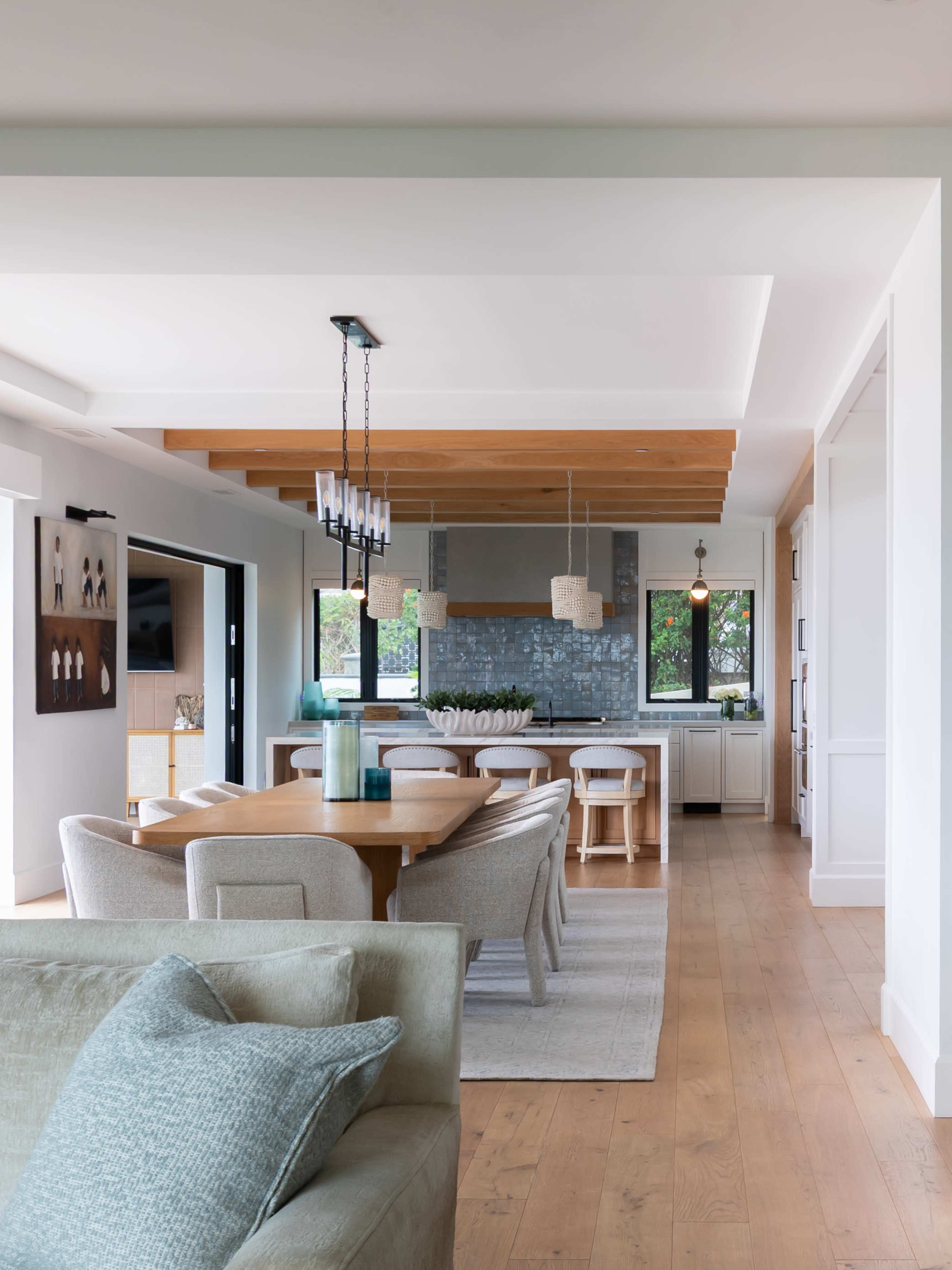A modern open-concept living and dining area features a wooden table, upholstered chairs, and a kitchen with stone backsplash and pendant lighting.