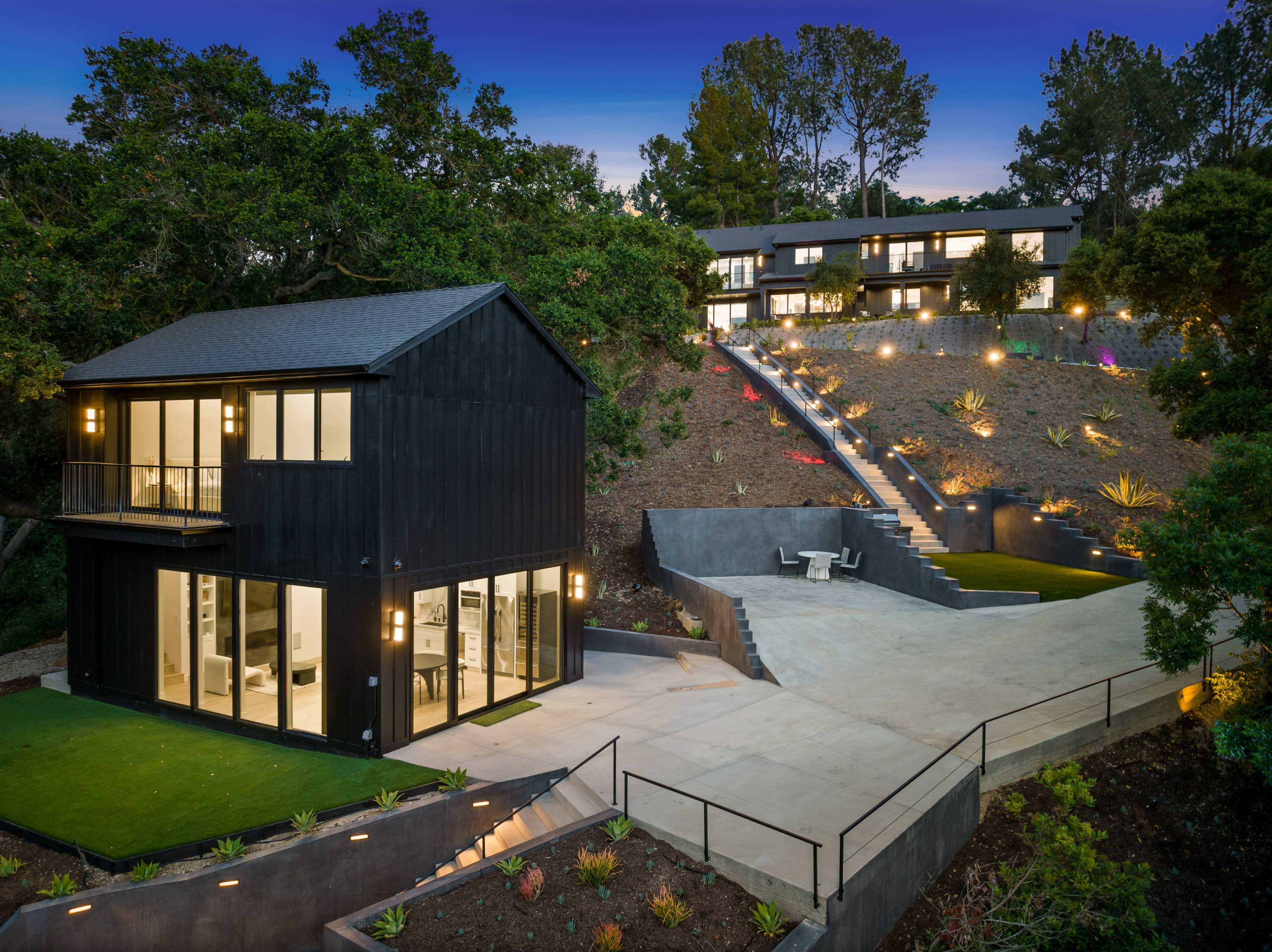 A modern black-framed house is situated in the foreground with a landscaped driveway leading to an elevated multi-level residence in the background, under a twilight sky.