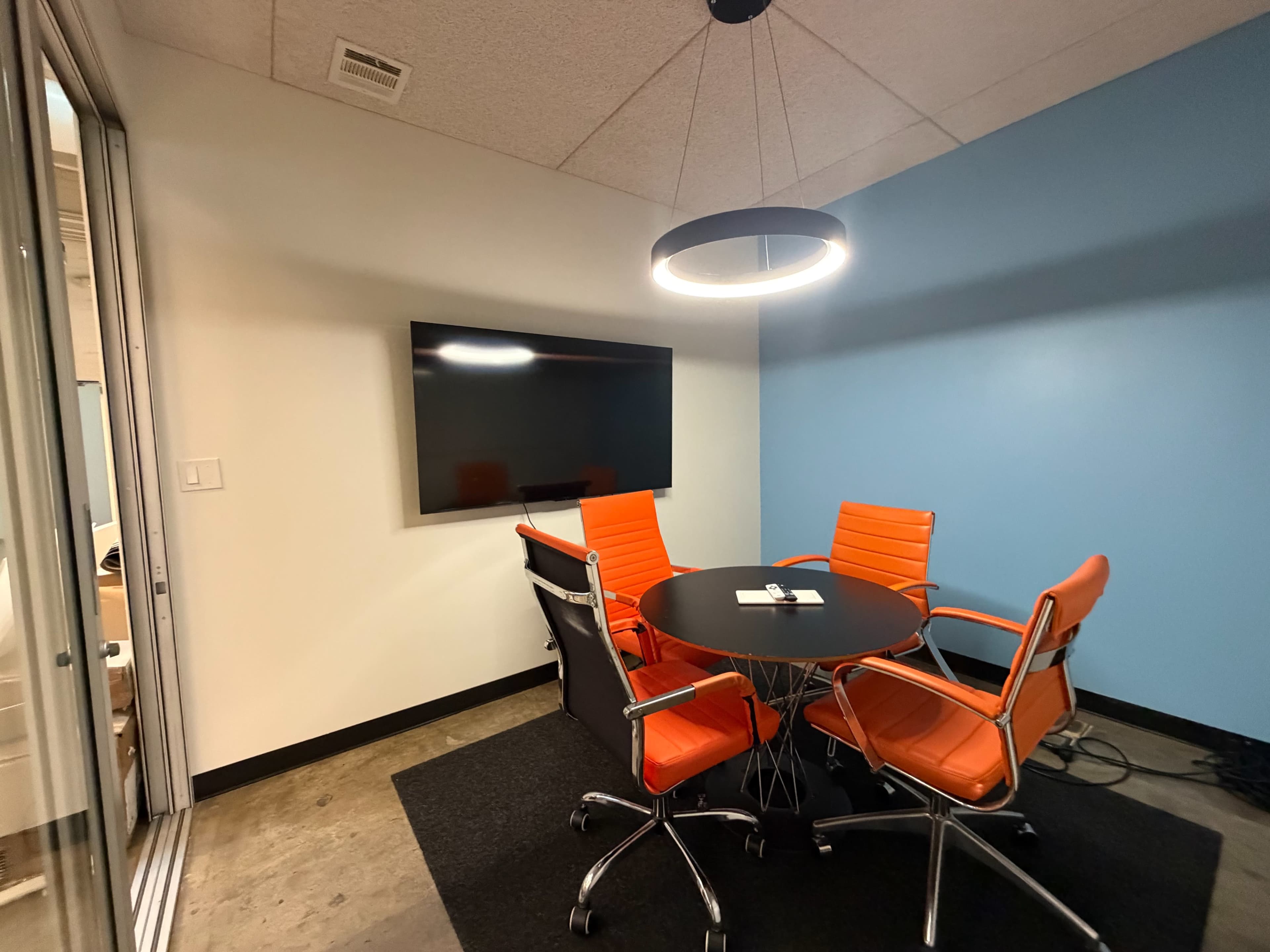 The image shows a small conference room featuring a round black table surrounded by four orange and black chairs, with a large wall-mounted television and a modern light fixture overhead.
