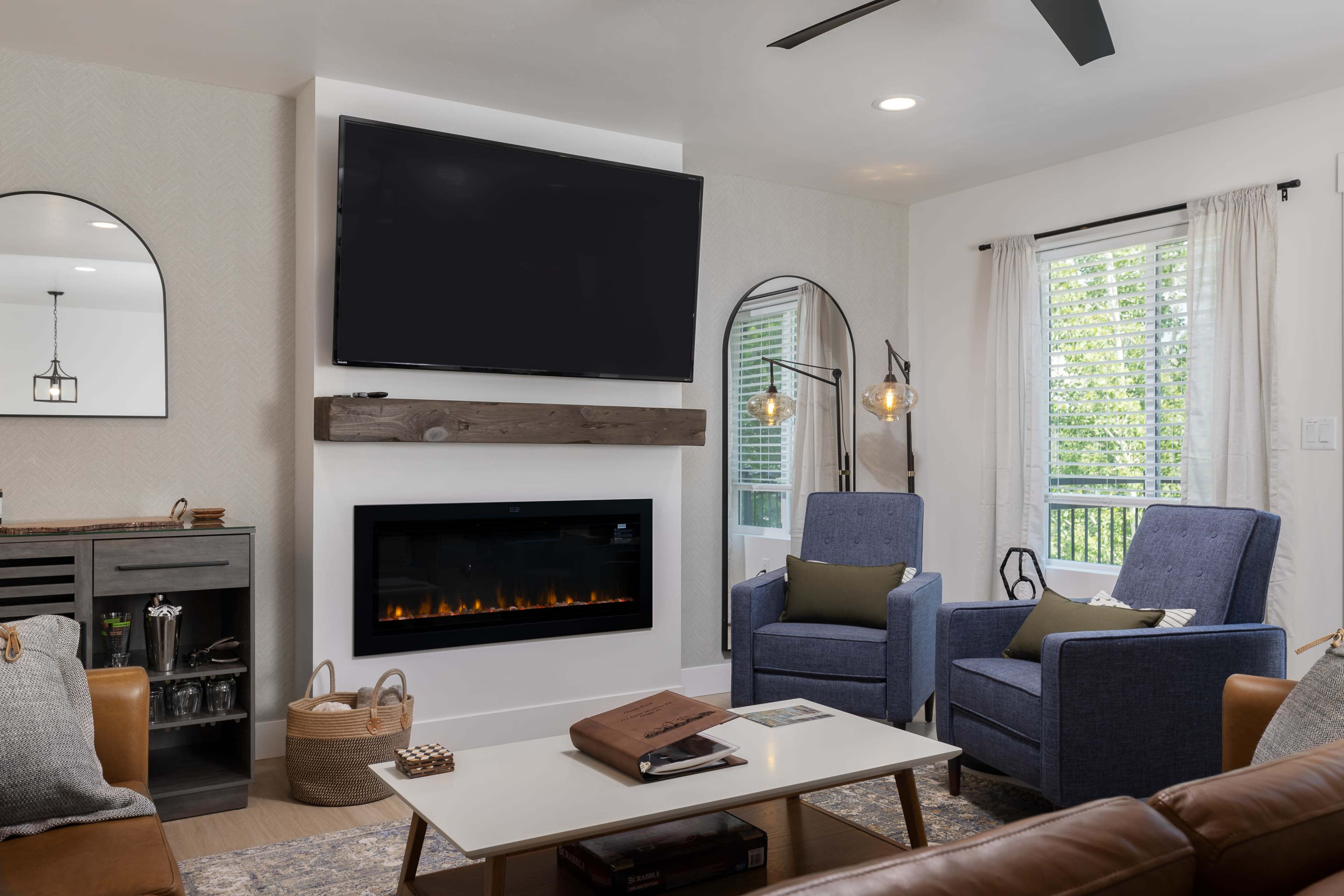 The living room features two blue armchairs facing a modern electric fireplace beneath a wall-mounted television, with windows providing natural light.