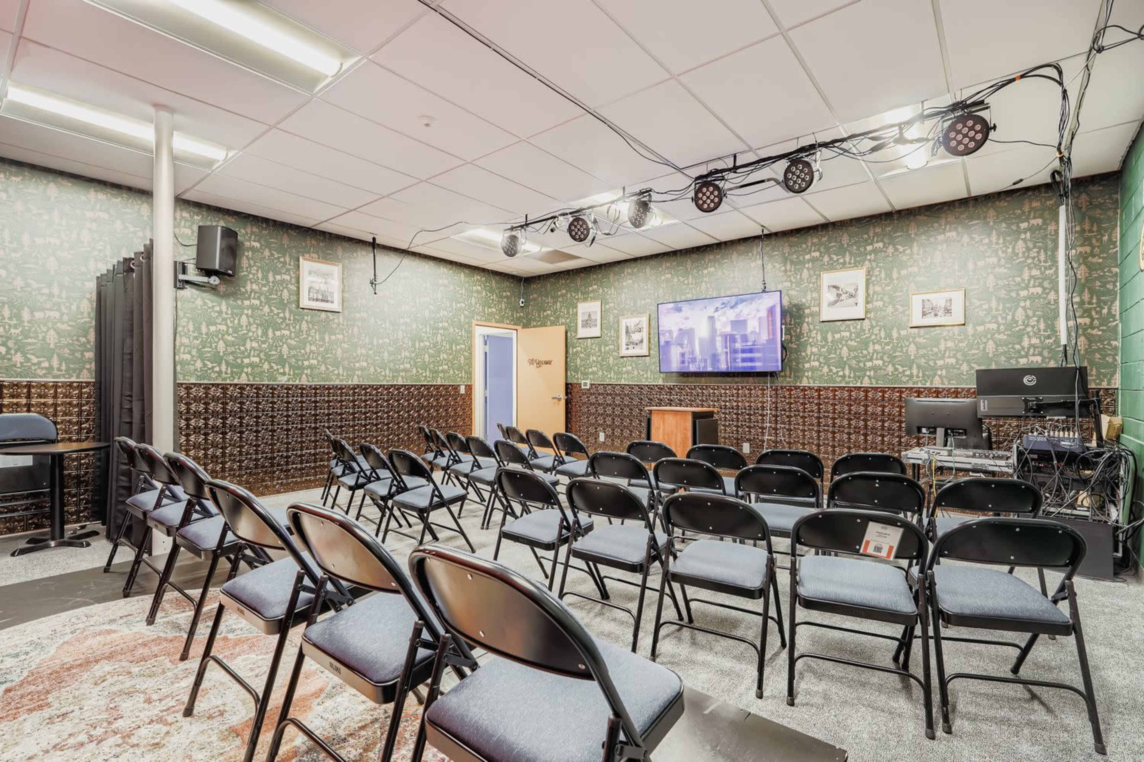 The image shows a small auditorium with rows of black folding chairs arranged facing a screen, against a textured green and brown wall.