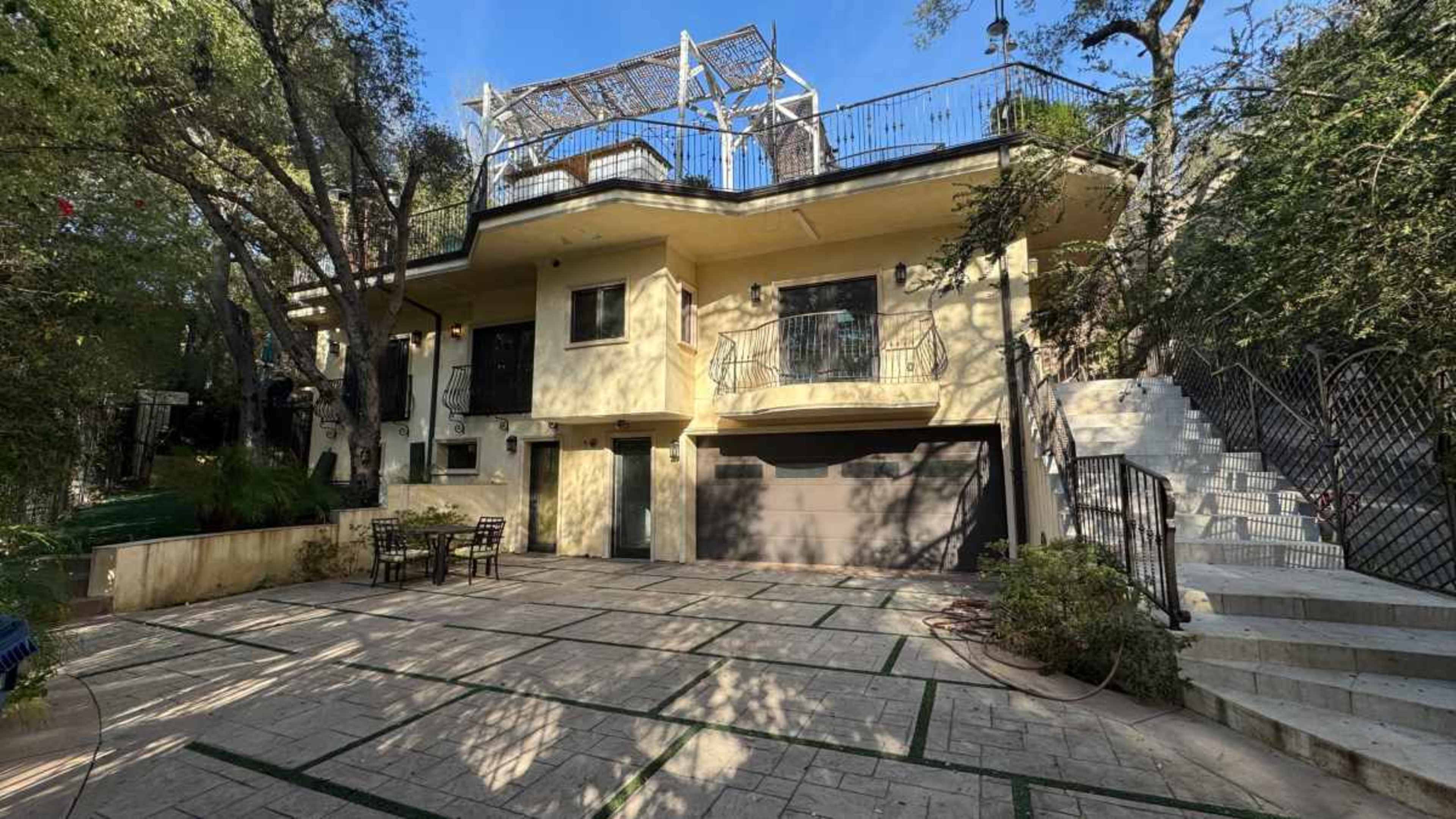 The image depicts a two-story house with a garage, surrounded by trees and featuring a rooftop terrace, along with a patio area with outdoor furniture.