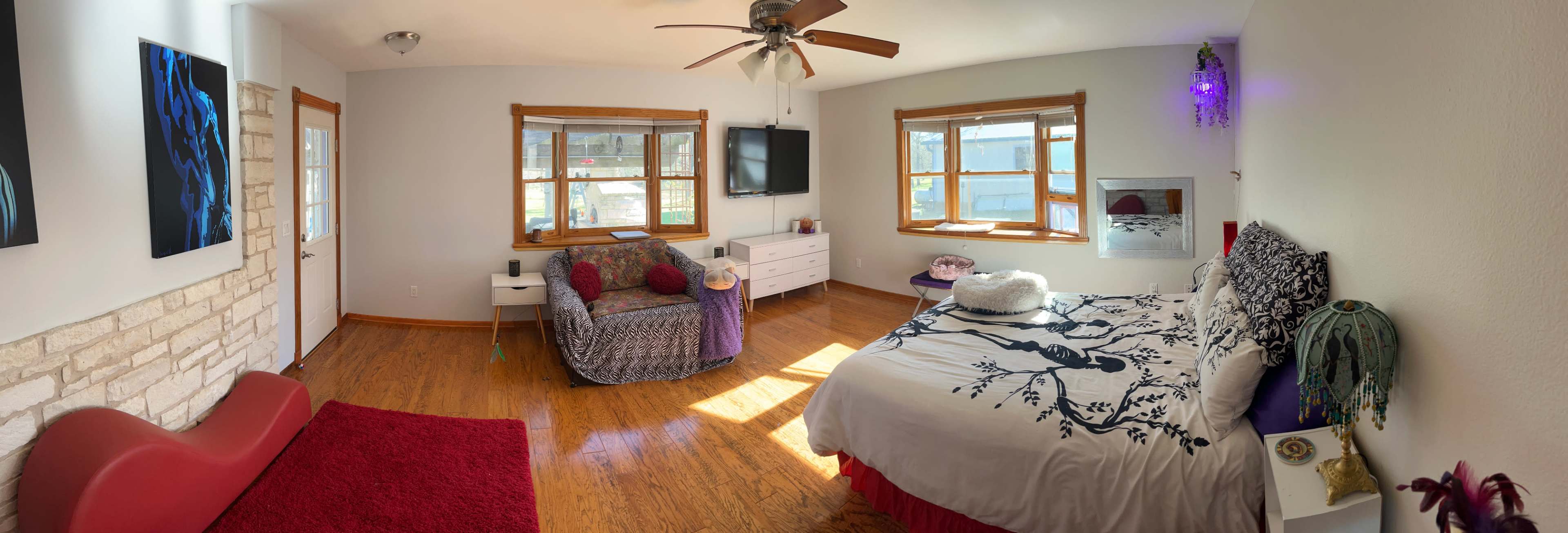 The image shows a spacious bedroom with a wooden floor, a bed covered in a patterned comforter, a chair with colorful throw pillows, and several windows allowing natural light to enter.
