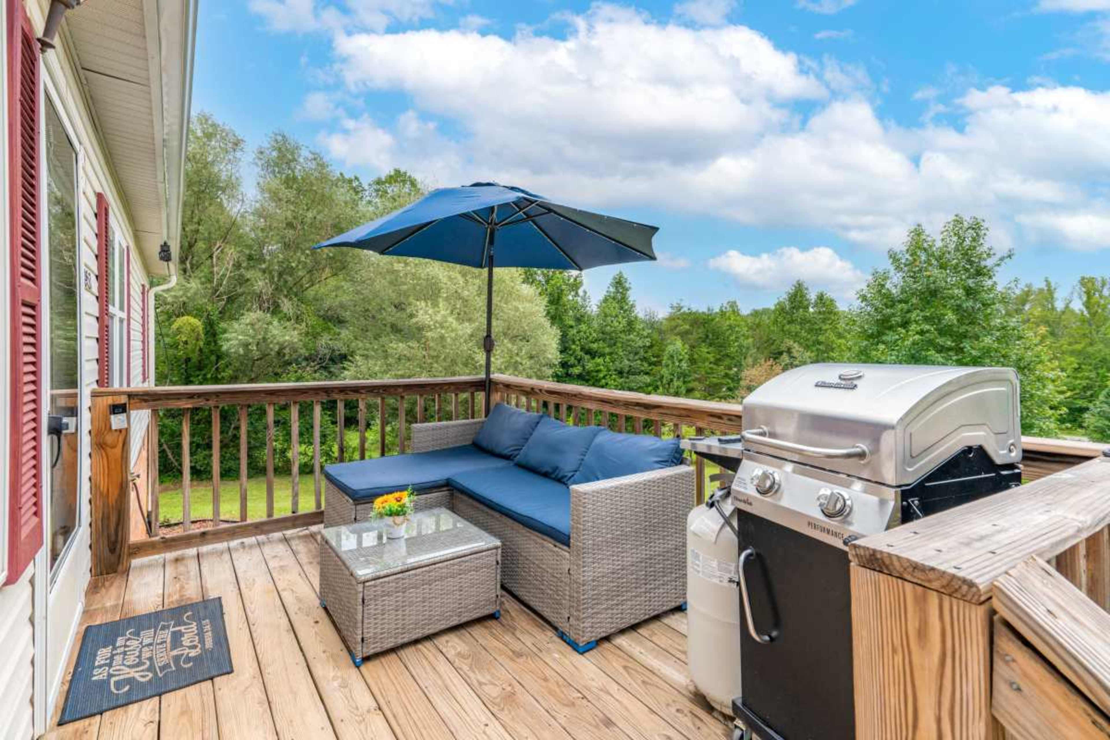 A wooden deck features a seating area with a blue sofa and umbrella, alongside a grill and natural greenery in the background.