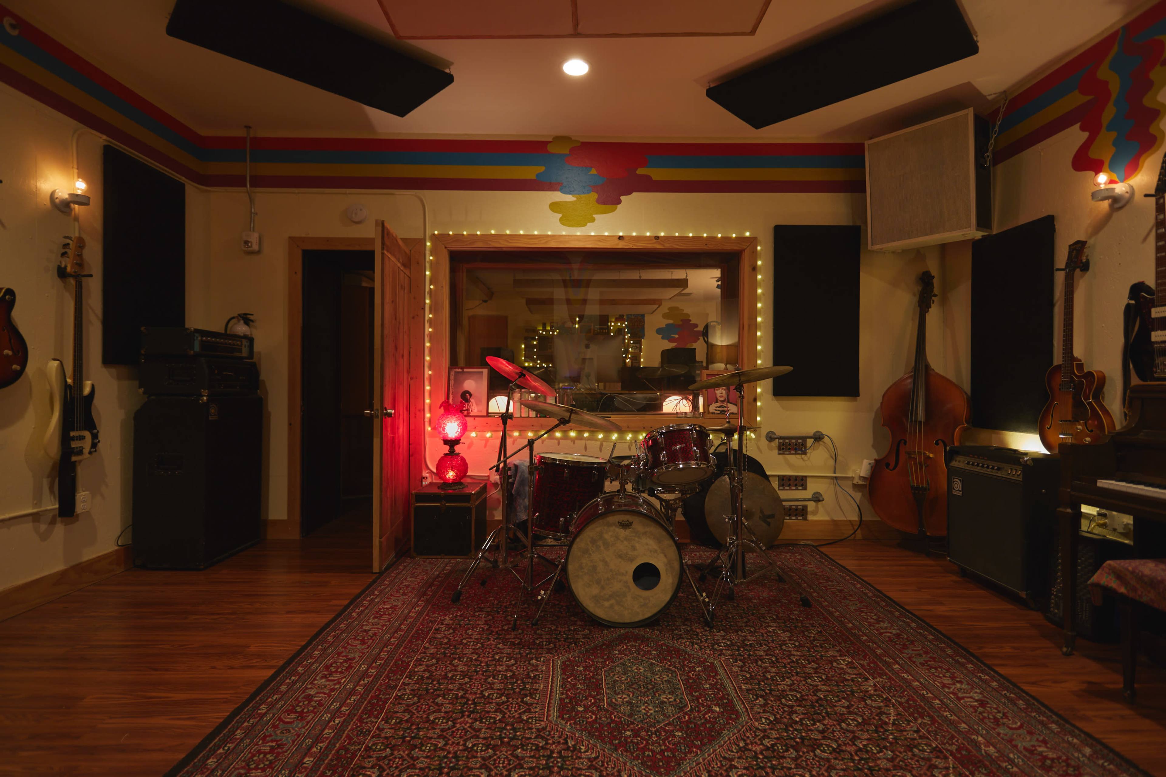 The image shows a music recording studio featuring a drum set in the center, surrounded by various musical instruments and equipment, with decorative lighting and acoustic panels on the walls.