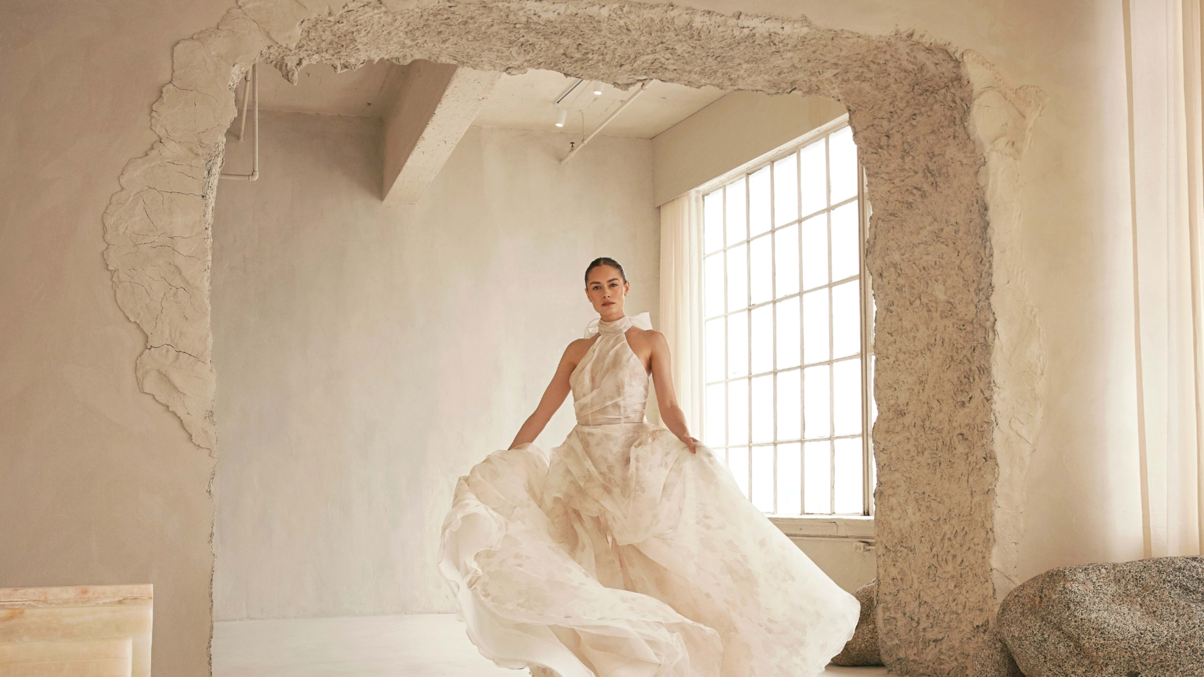A woman in a flowing, light-colored dress stands gracefully in a modern, minimalist room with a large window and textured wall.