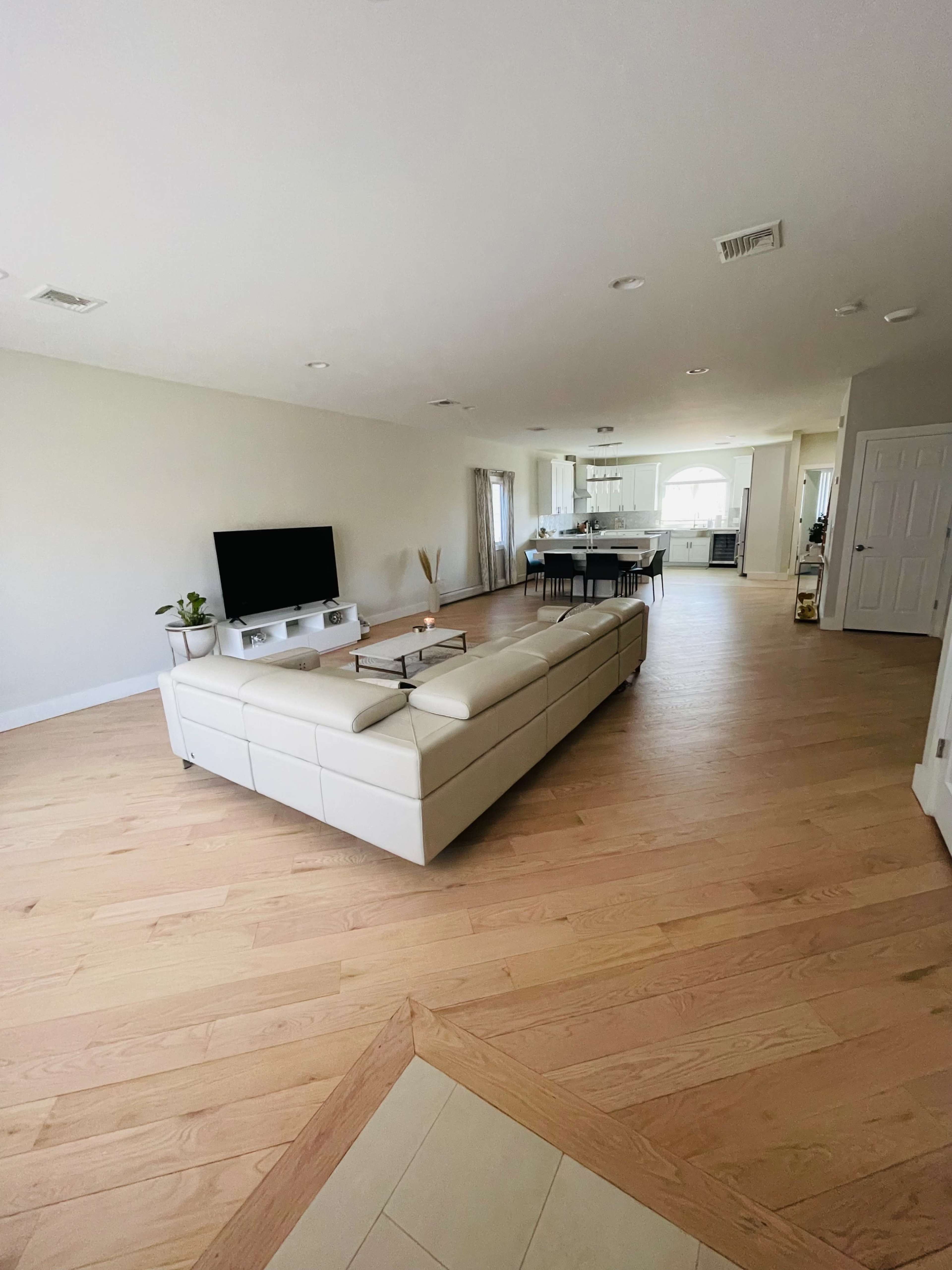 A spacious, modern living room features a white sectional sofa and a television, overlooking an open kitchen area with large windows.