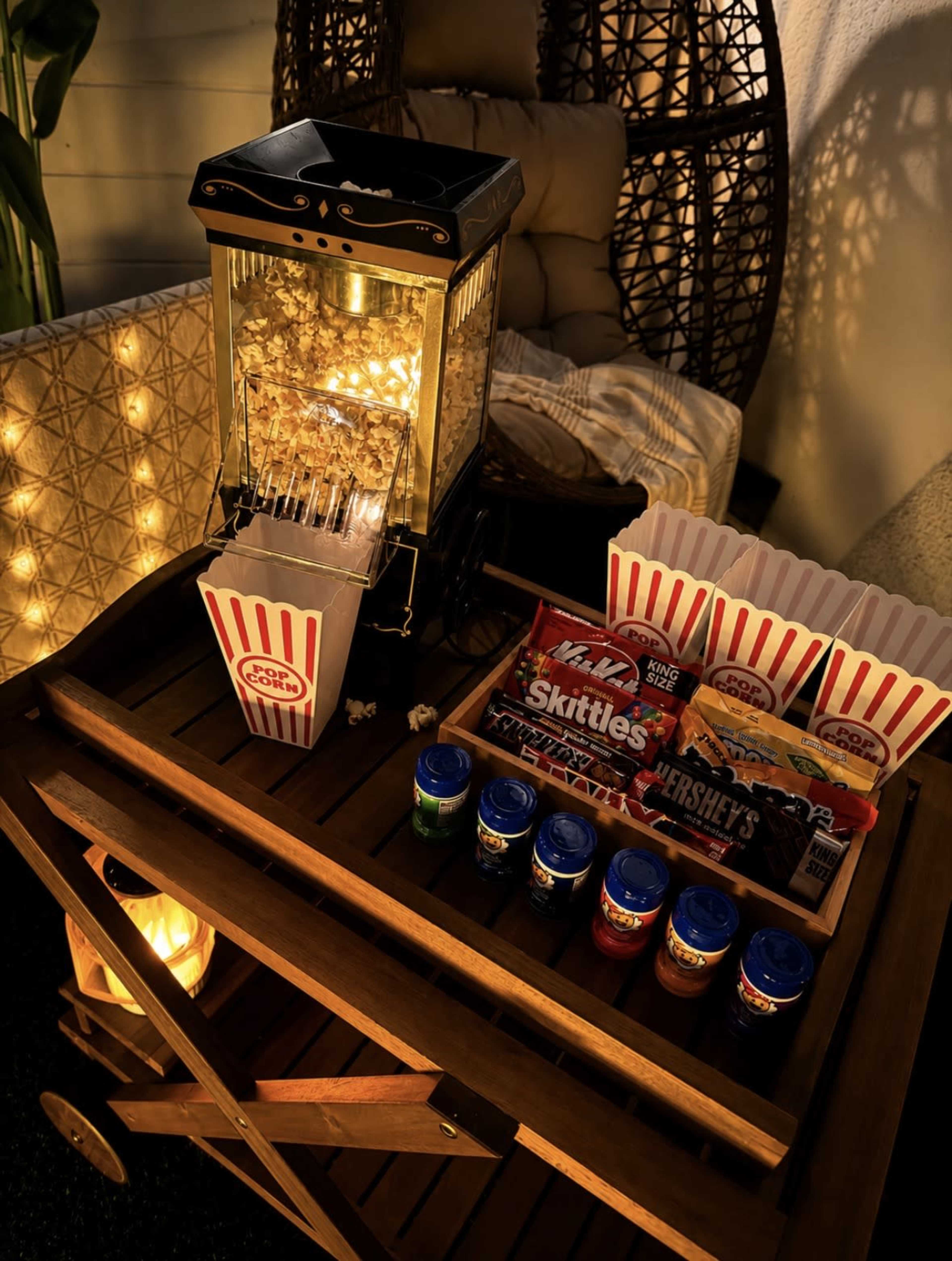 A popcorn machine sits on a wooden cart next to various candies and snacks, illuminated by warm lights.