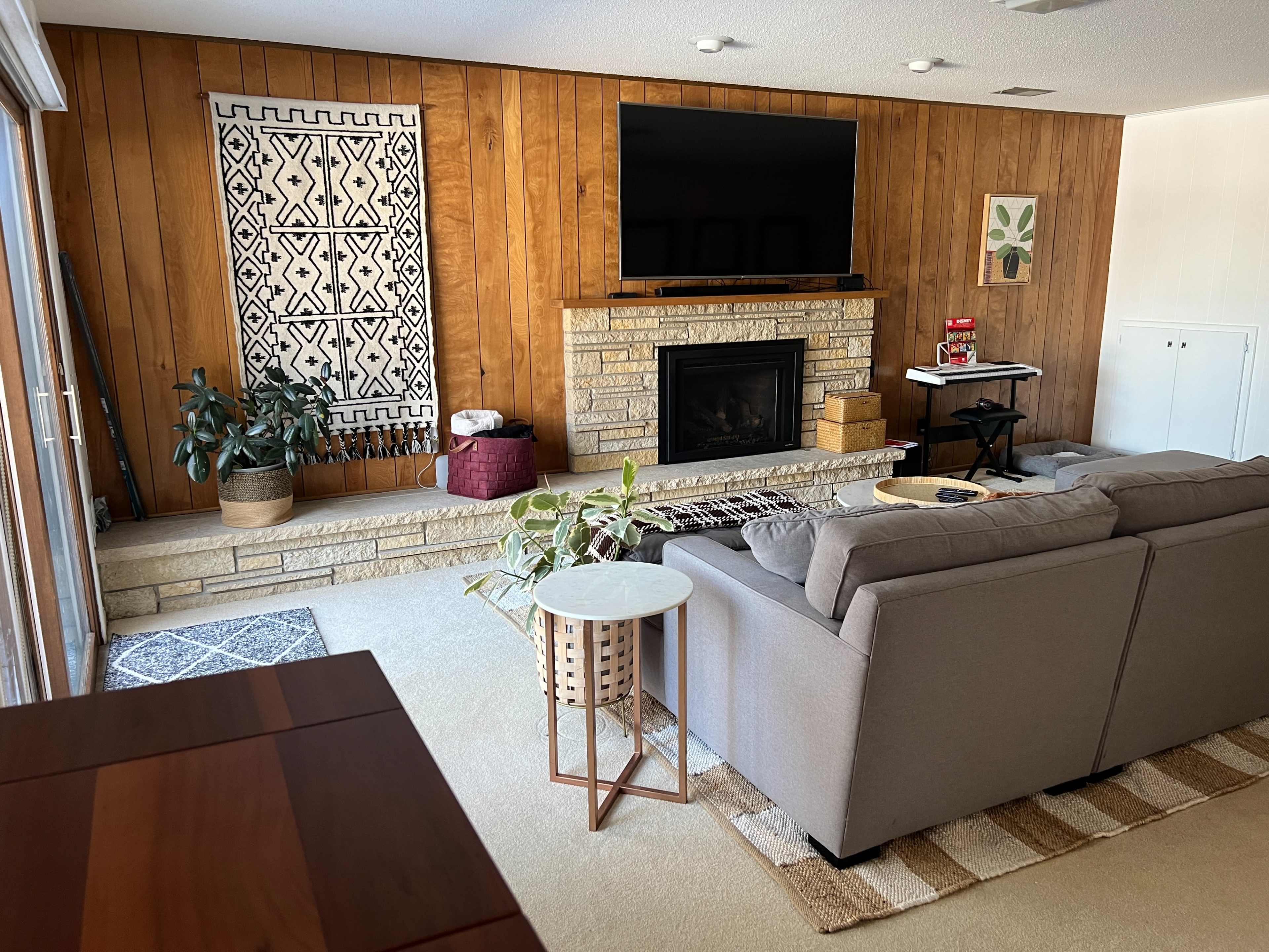 A living room with a grey sofa, a stone fireplace, a television mounted on the wall, and a wooden wall adorned with decorative art.