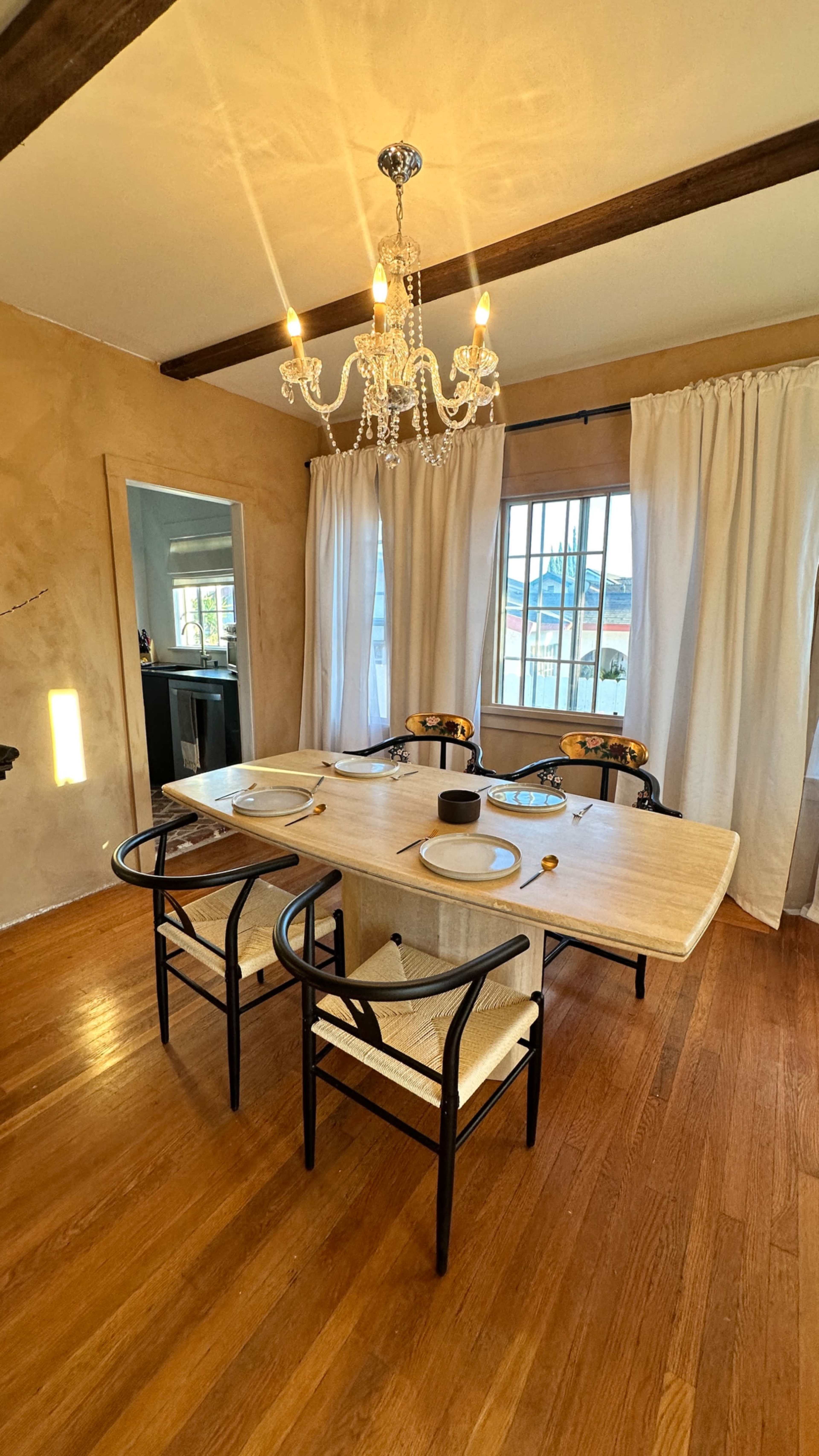 A modern dining room features a light wooden table surrounded by four chairs, illuminated by a chandelier with a warm glow.