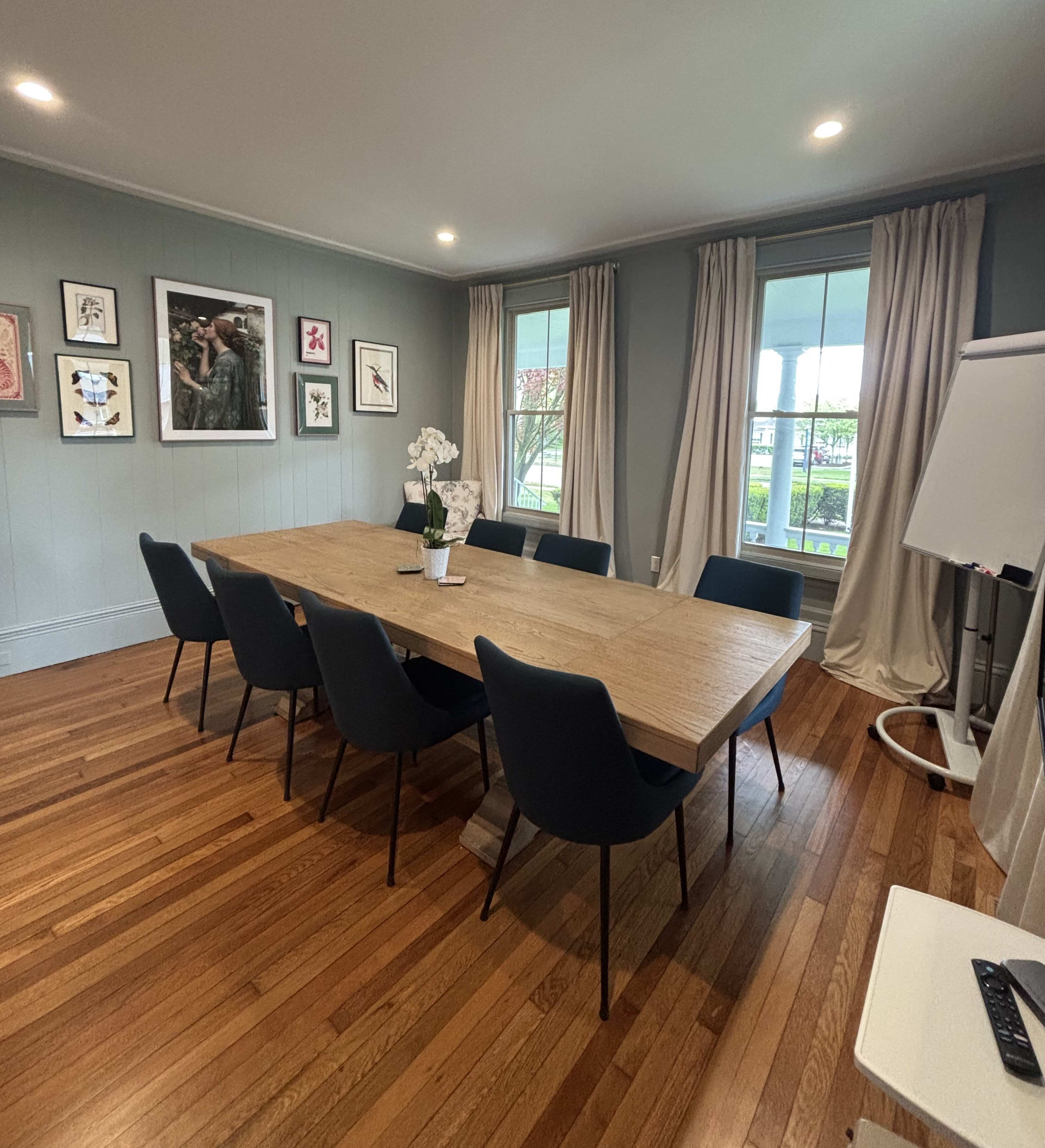 The image shows a modern conference room with a large wooden table surrounded by black chairs, framed artwork on the walls, and curtains covering the windows.