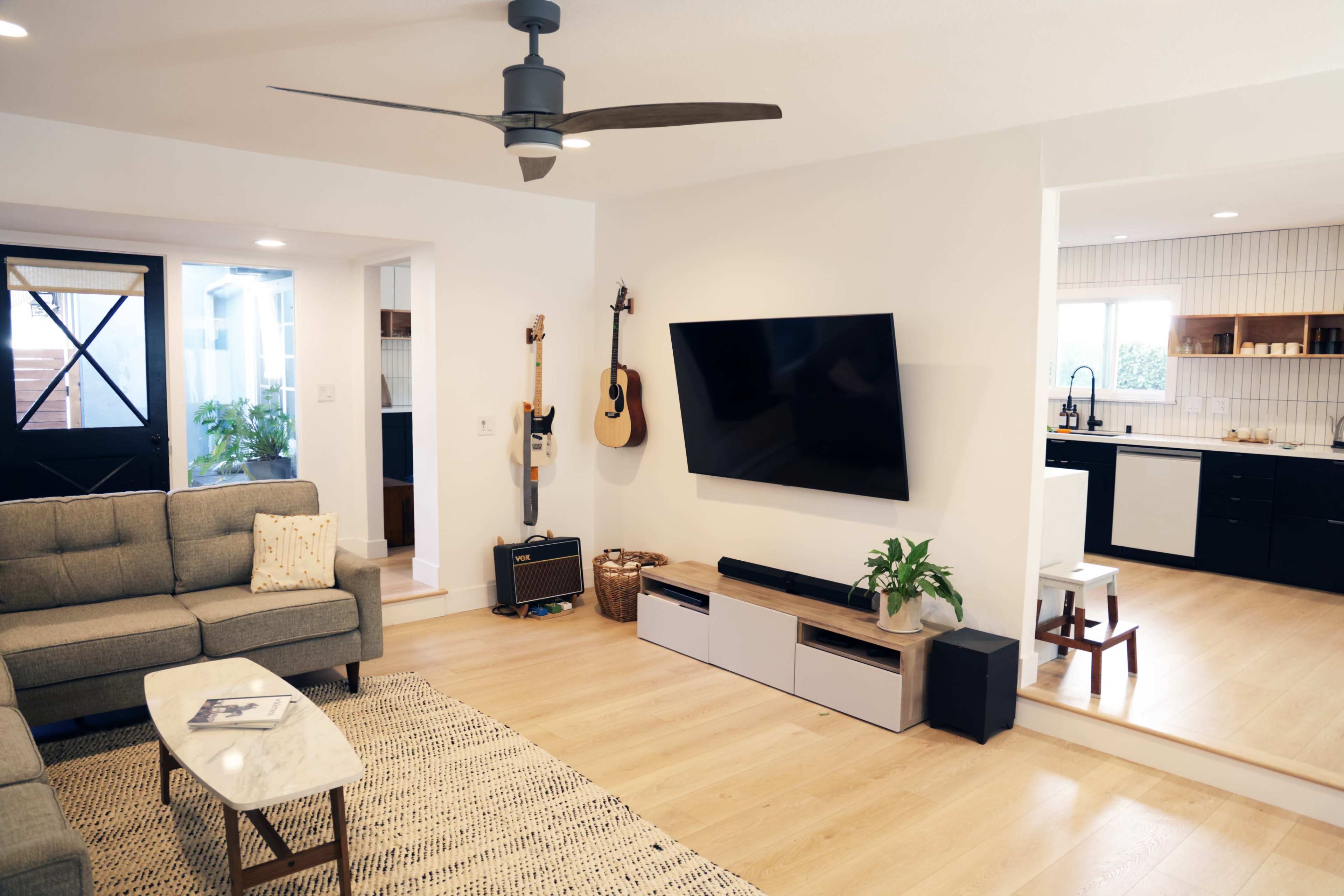 A modern living room with a gray sofa, a mounted flat-screen TV, a guitar on display, and a connected kitchen area featuring white cabinets and a wooden floor.