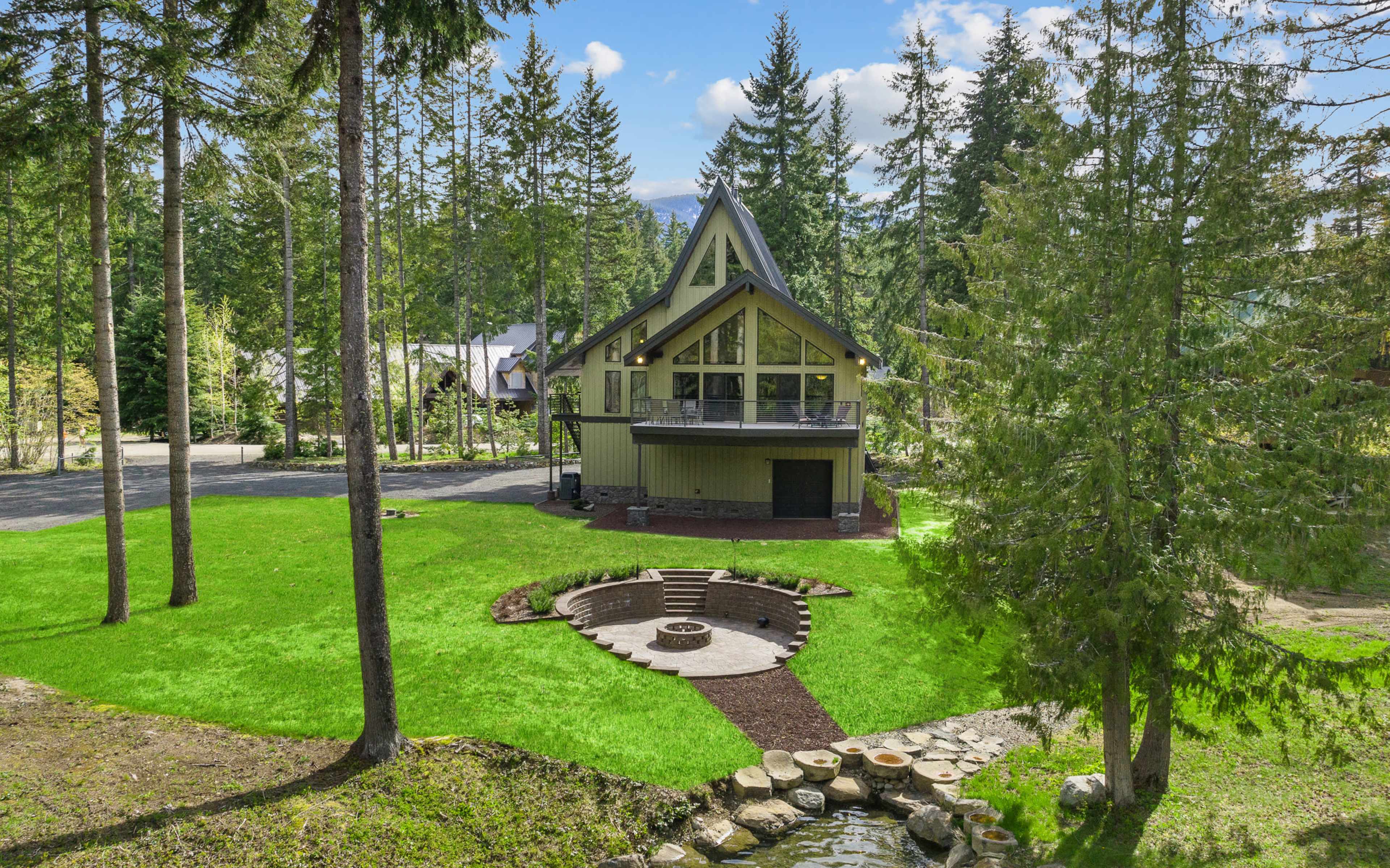 A three-story house with a distinctive architectural design is set in a landscaped garden featuring a circular stone seating area and surrounded by tall trees.