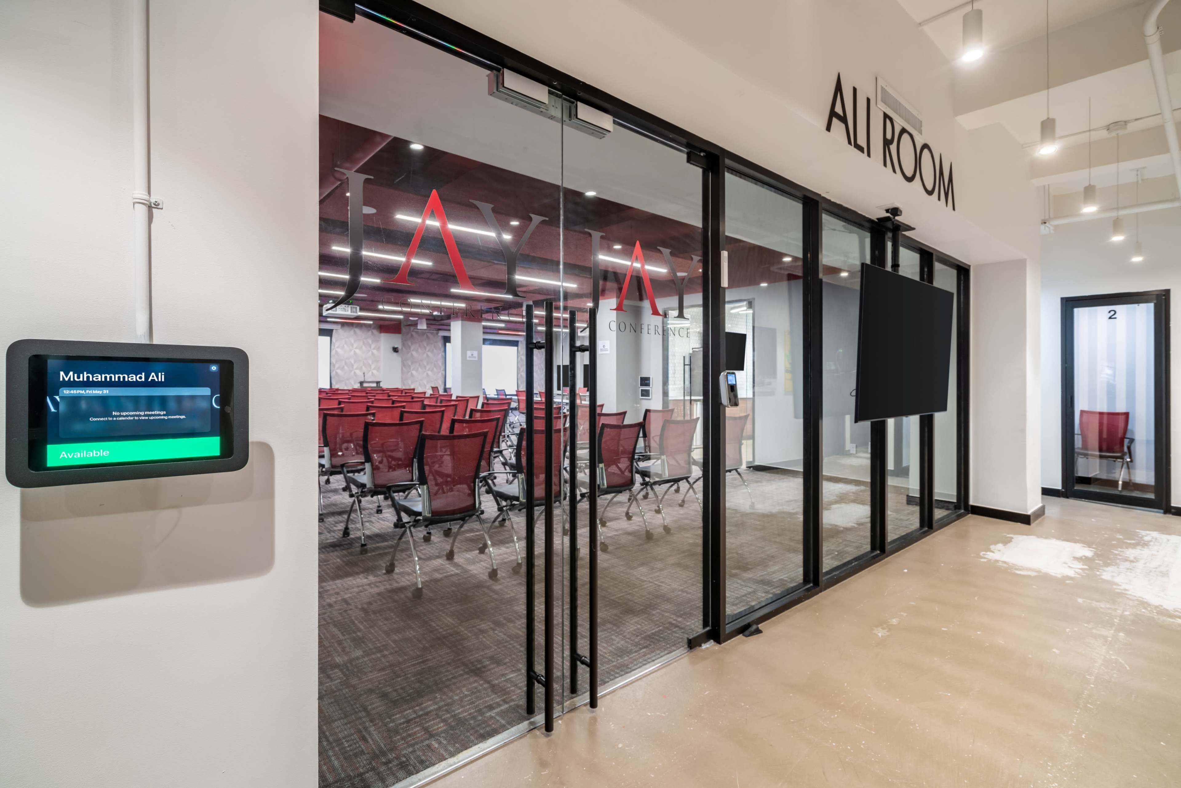 The image shows the entrance to a conference room named "Ali Room," featuring glass doors and a digital display indicating its availability.