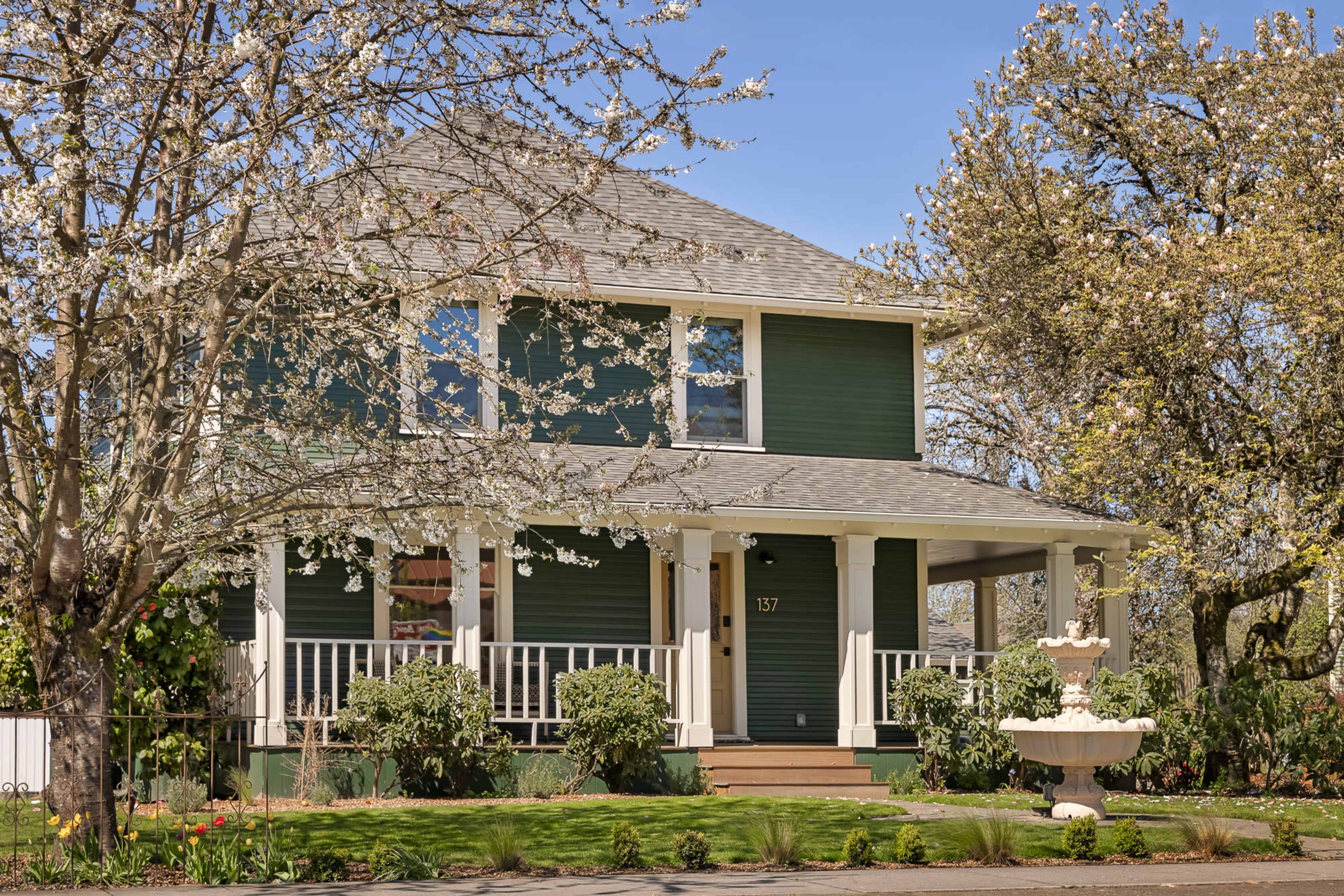 A two-story green house with a front porch is surrounded by blooming trees and a decorative fountain in the yard.