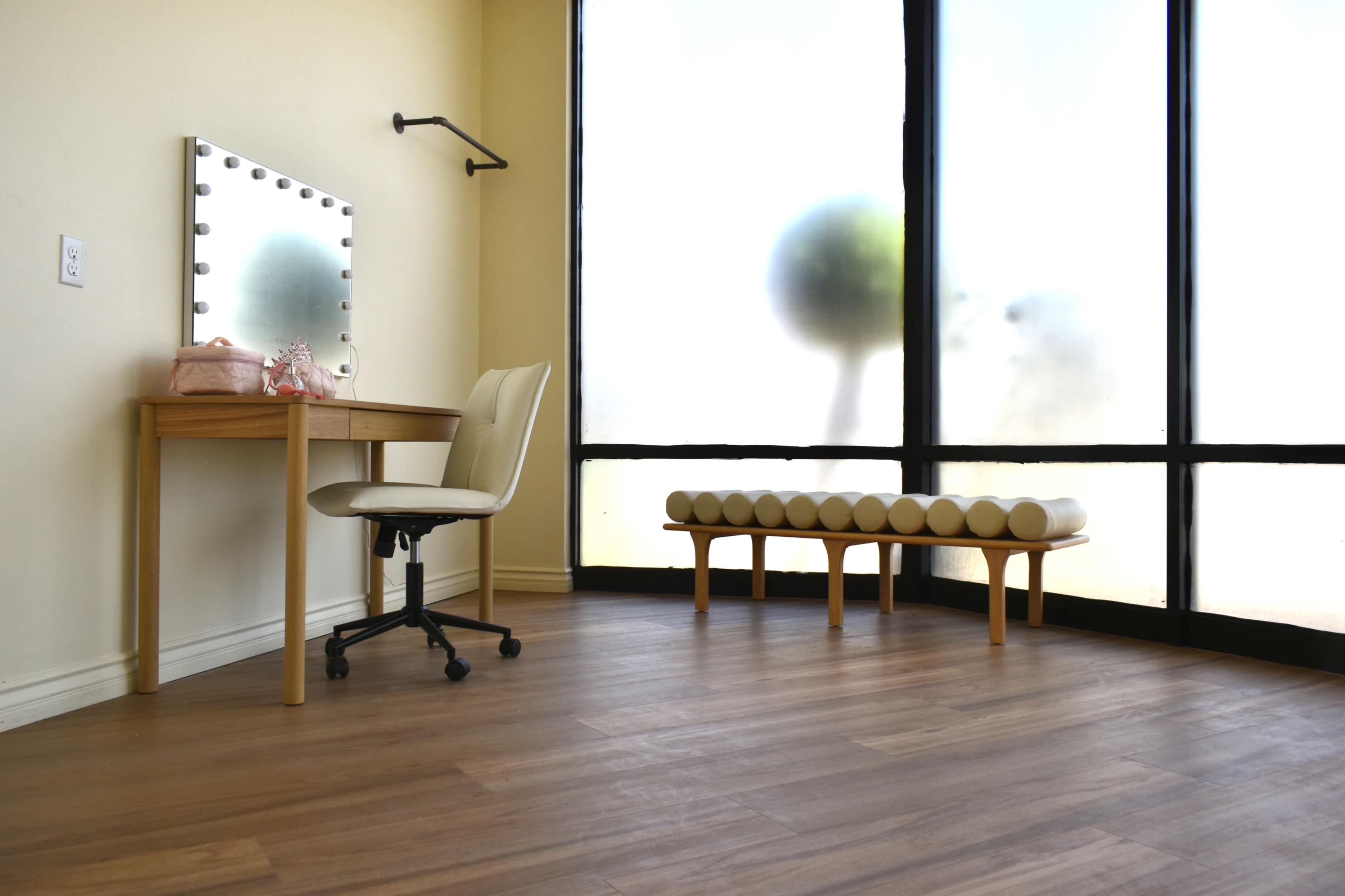 A well-lit room features a vanity table with a mirror and a chair, alongside a bench by a large, frosted glass window.