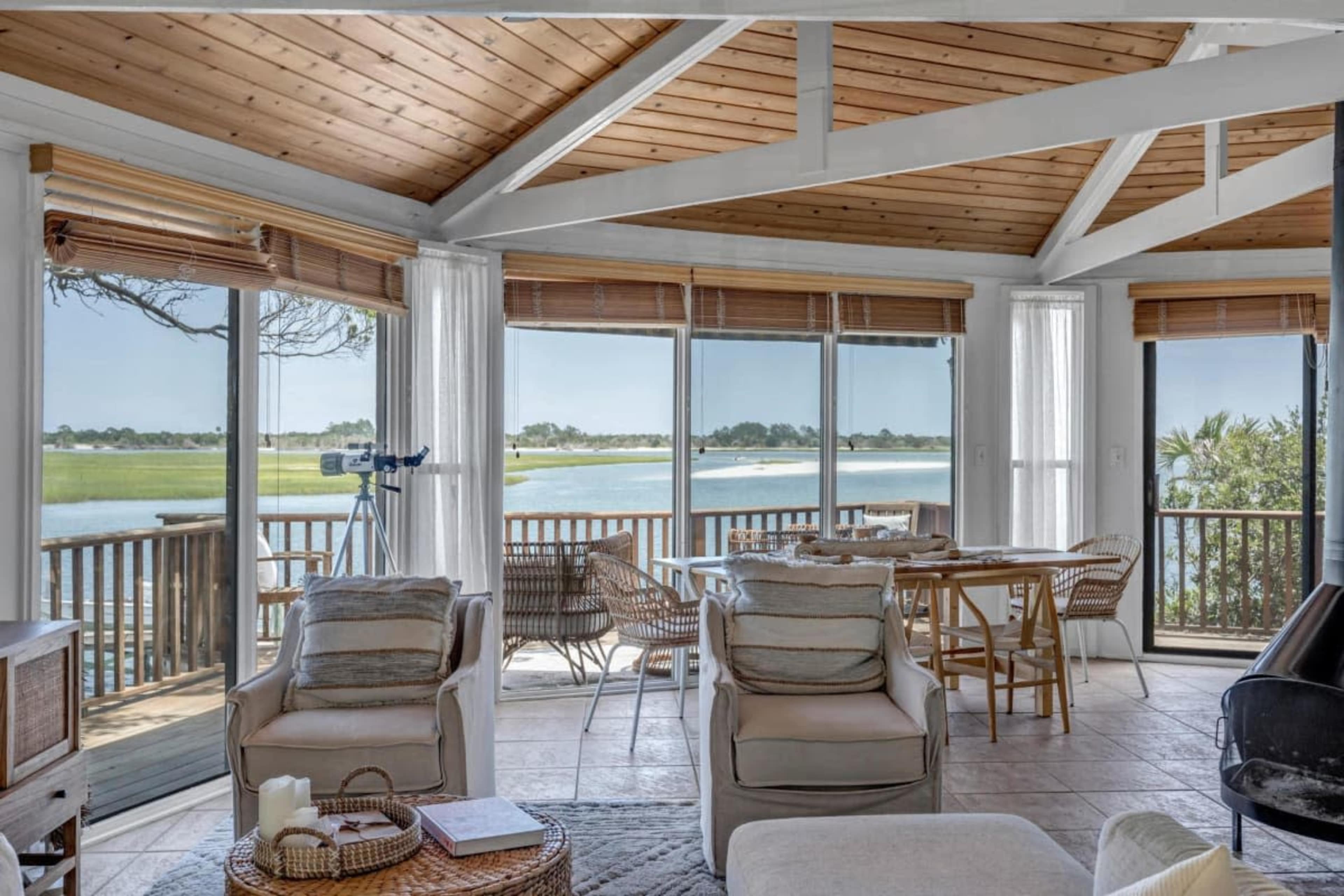 A bright, open room features large windows overlooking a water view, with a dining area and comfortable seating arrangements.