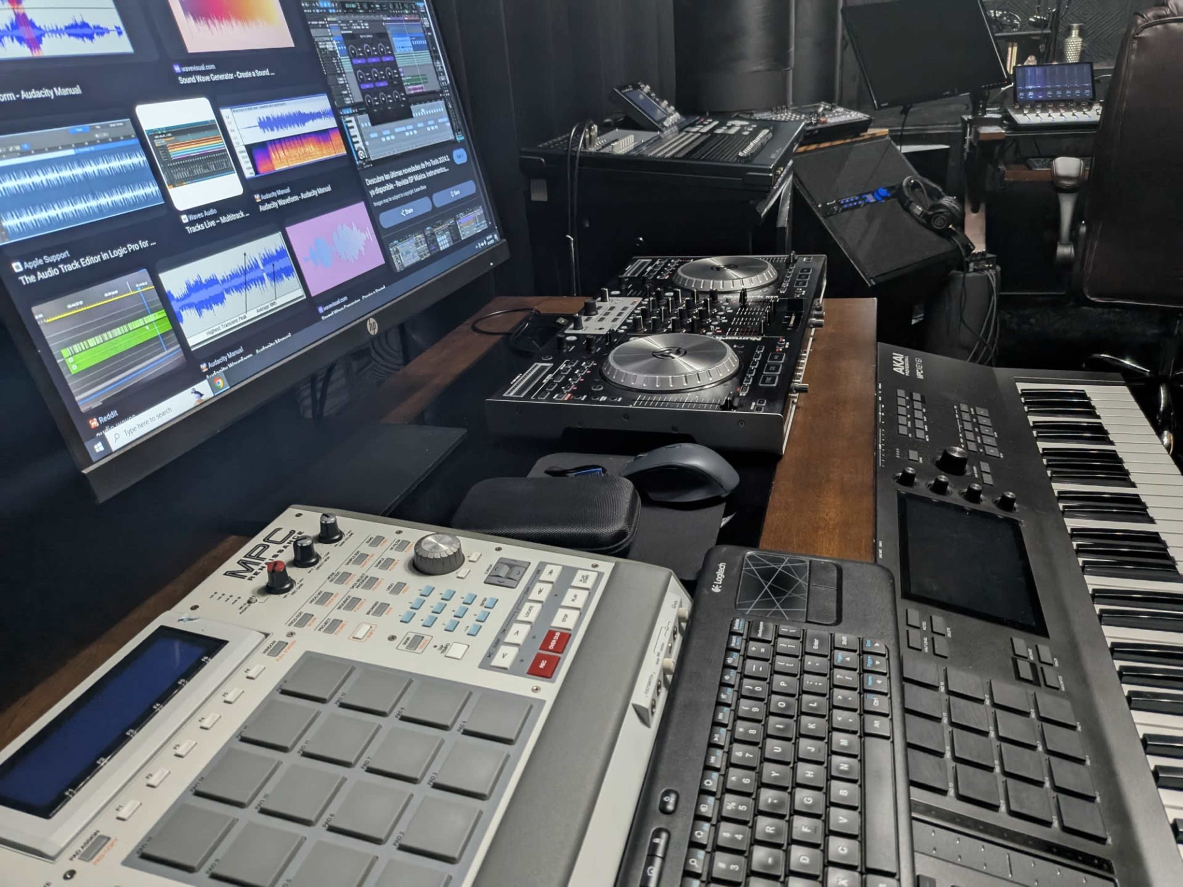 The image shows a music production setup featuring a computer monitor with audio software, a DJ controller, a MIDI keyboard, and a drum machine.