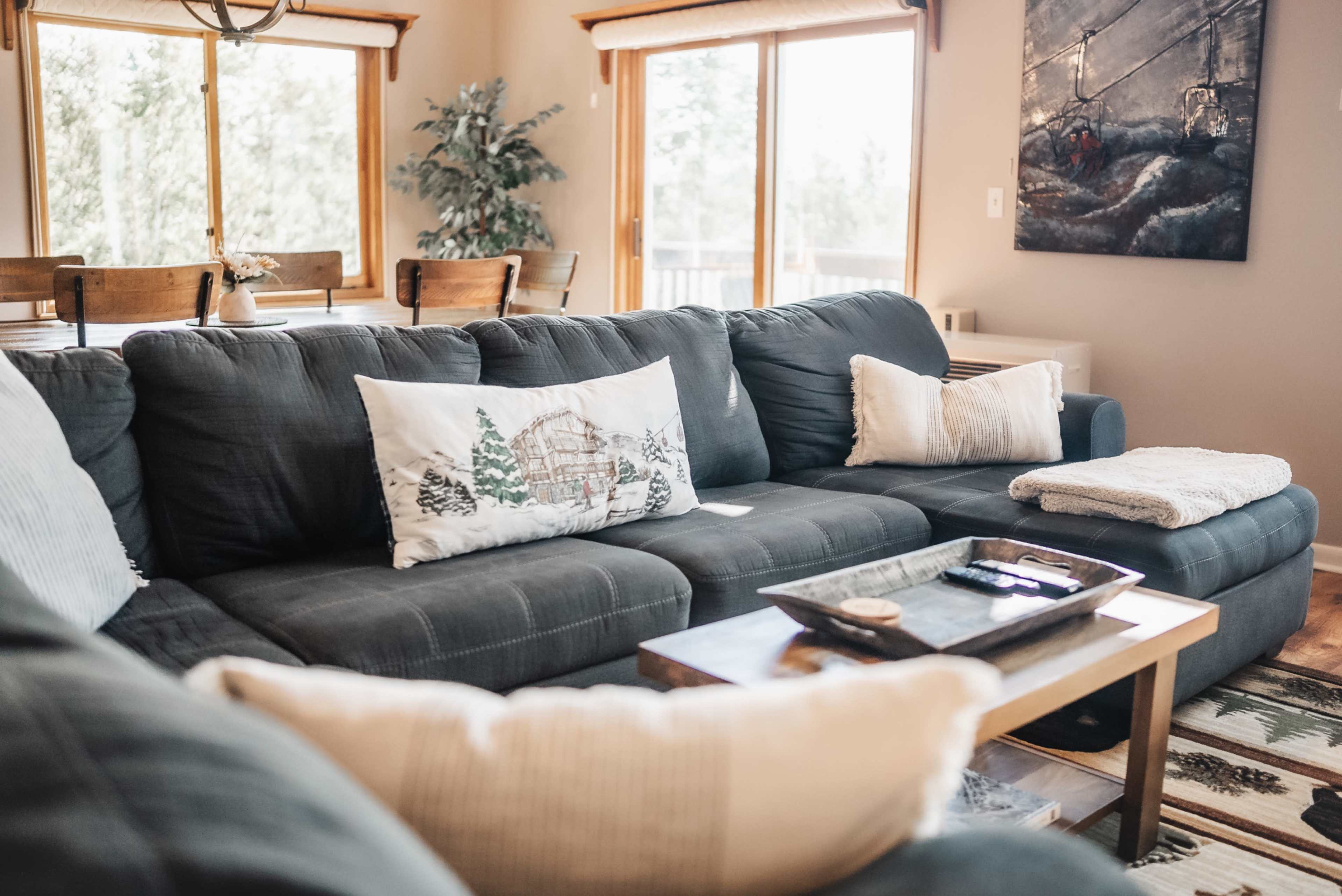 A spacious living room features a dark gray sectional sofa with decorative pillows, a wooden coffee table, and large windows that allow natural light to fill the space.