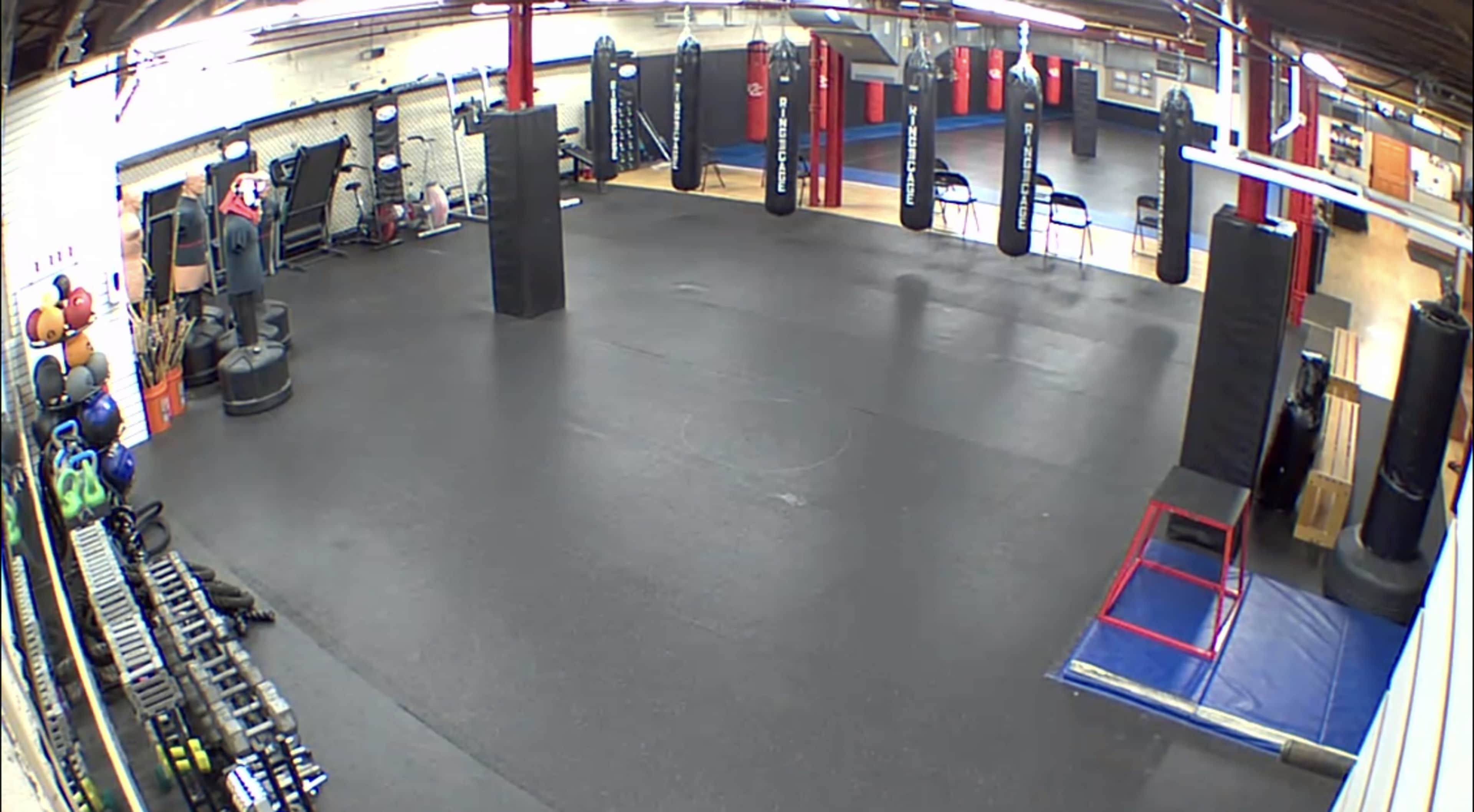 The image shows a spacious gym interior with various workout equipment, including punching bags, weights, and exercise mats arranged on a rubber floor.