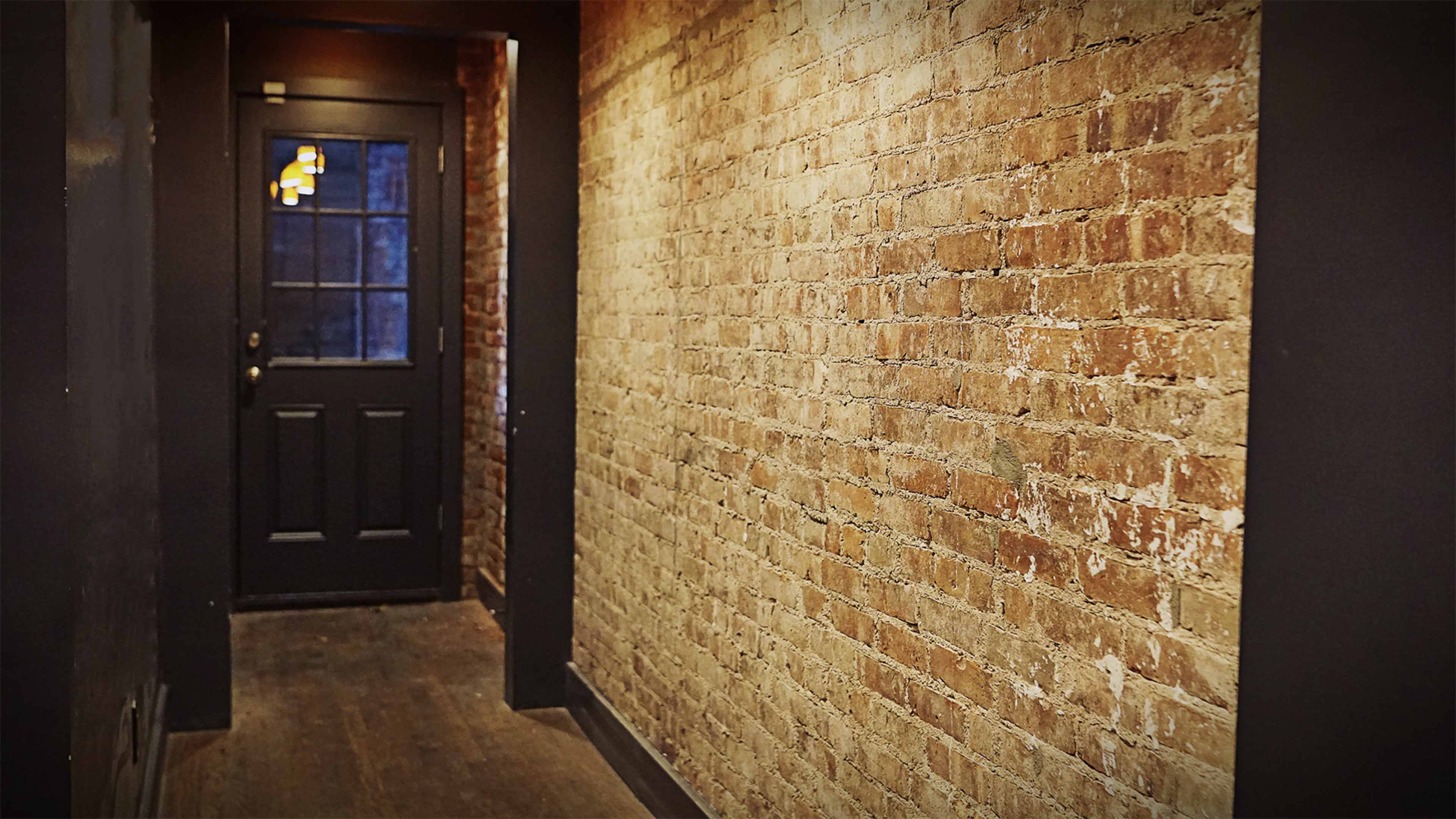 The image features a narrow hallway with exposed brick walls and a closed black door at one end.