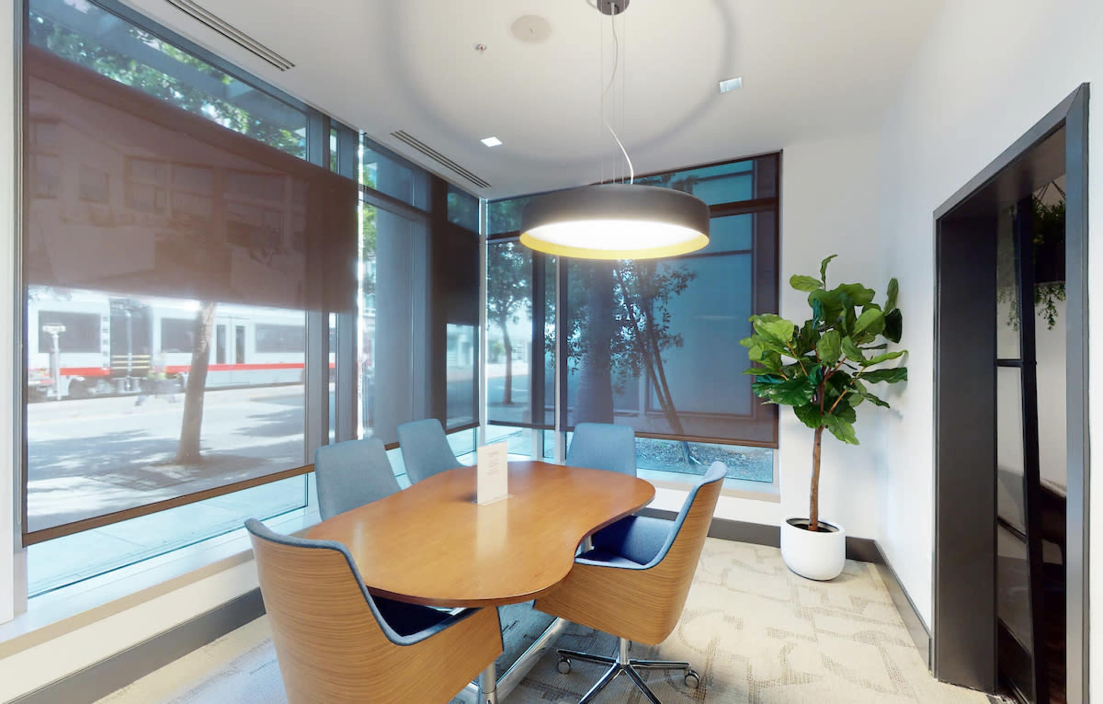A modern conference room with a wooden table and blue chairs, featuring large windows with shades and a decorative plant in the corner.