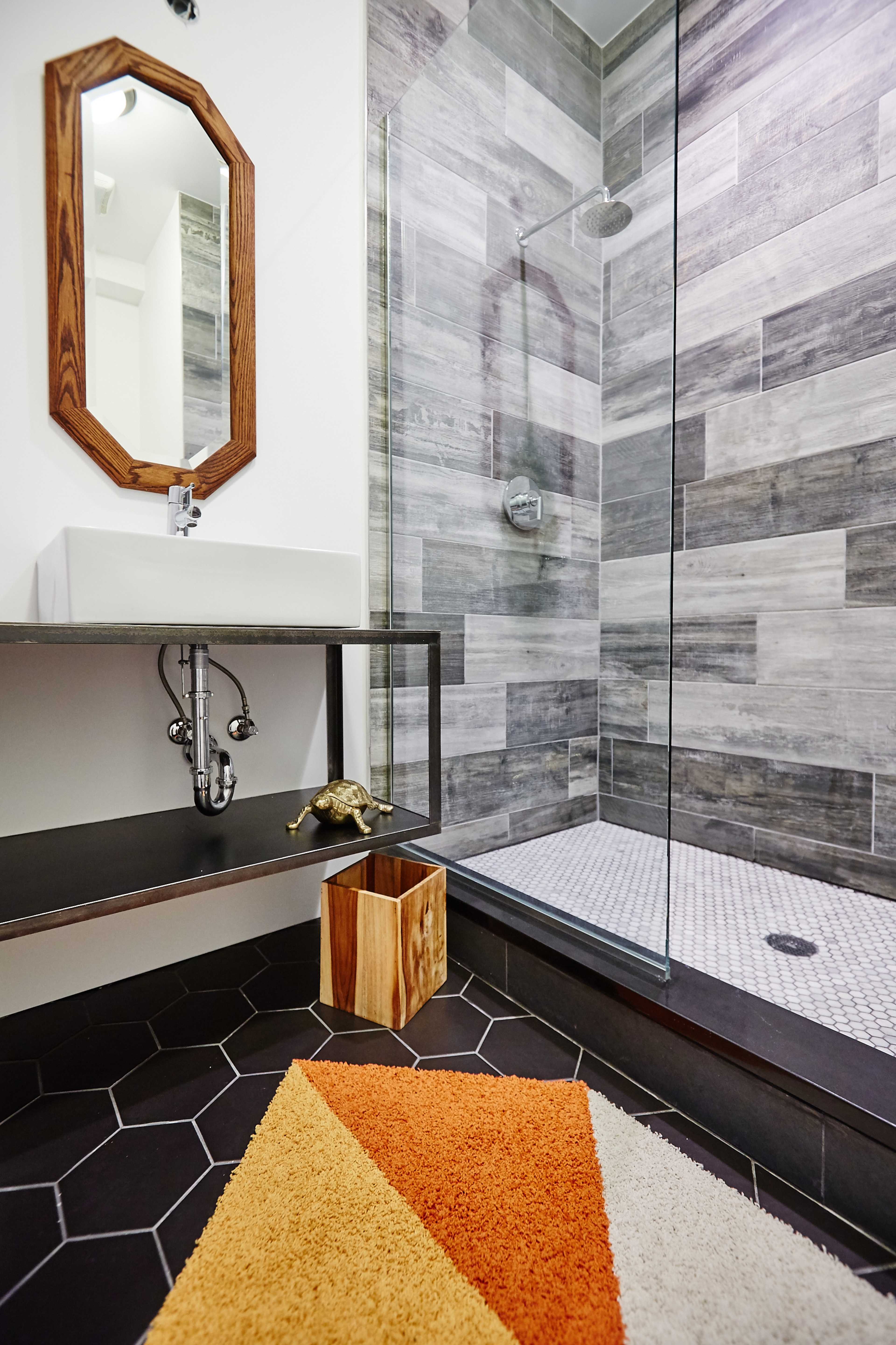 The image shows a modern bathroom featuring a glass shower, a sink with a wooden mirror frame, and a geometric rug on the floor.